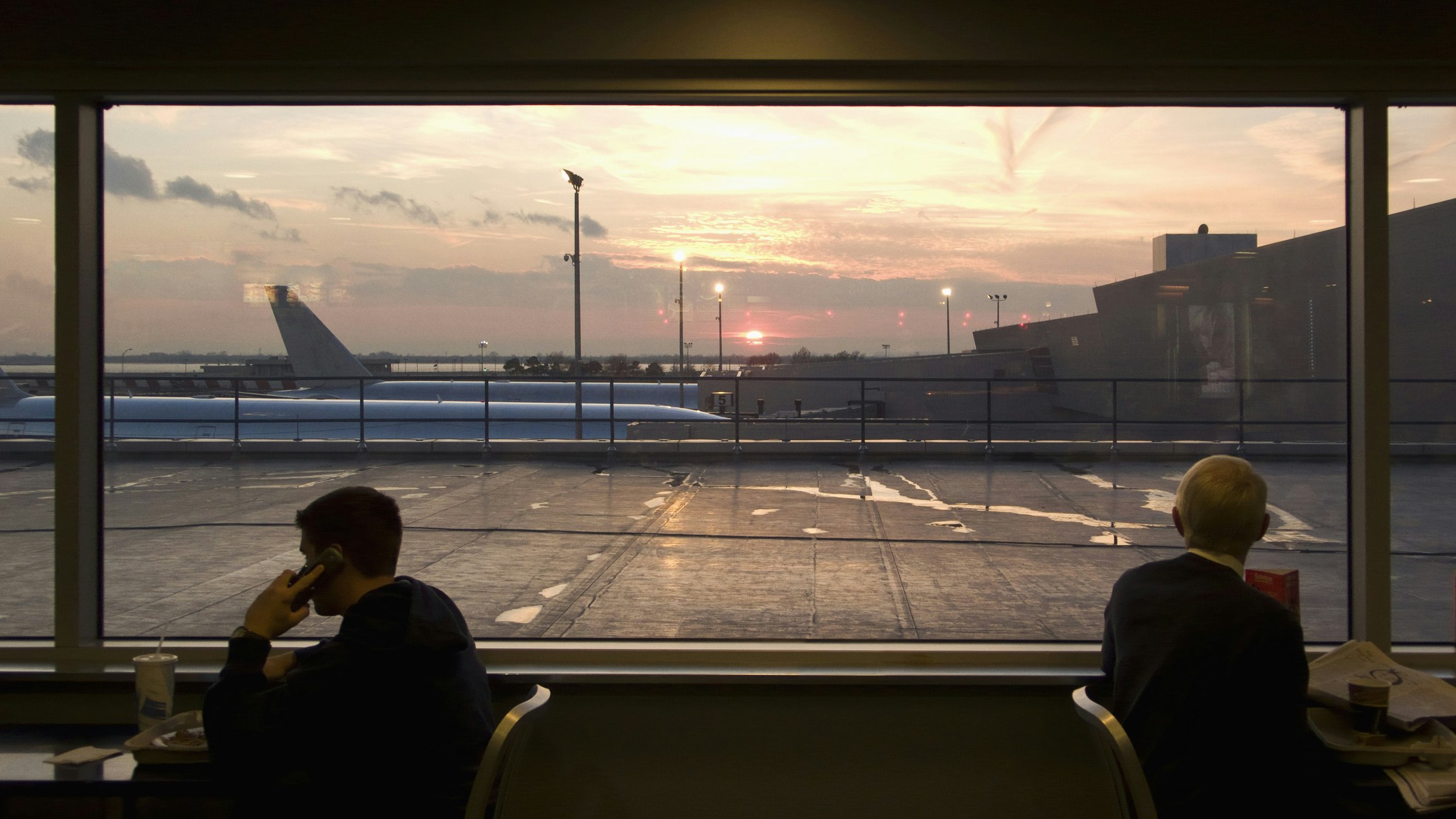 Two men, one younger and one older, sit in dark clothing almost silhouetted against the air terminal outside at sunrise
