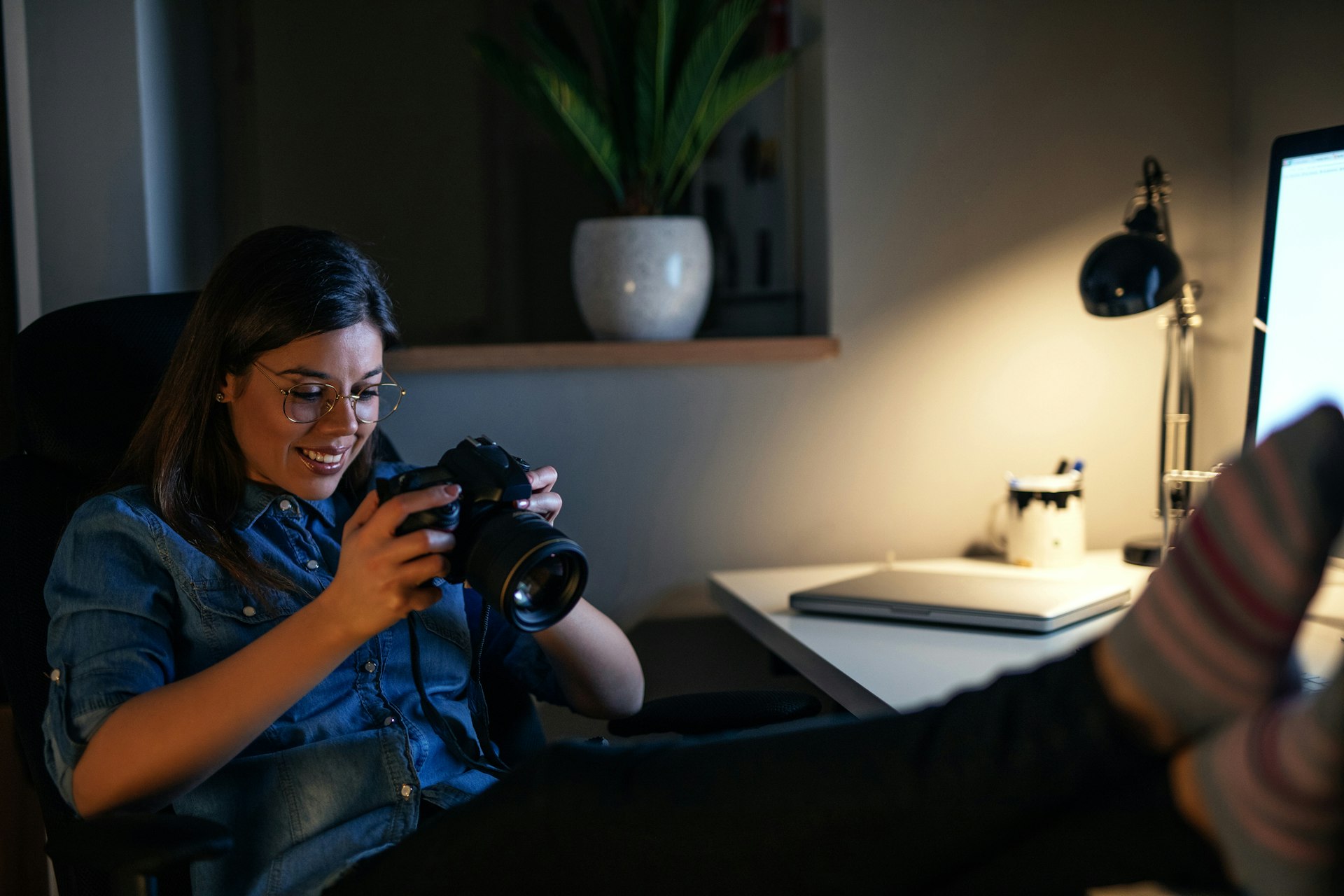 A woman in a blue chambray shirt and round glasses sits with her feet propped up on her desk looking at photos on her camera with a smile