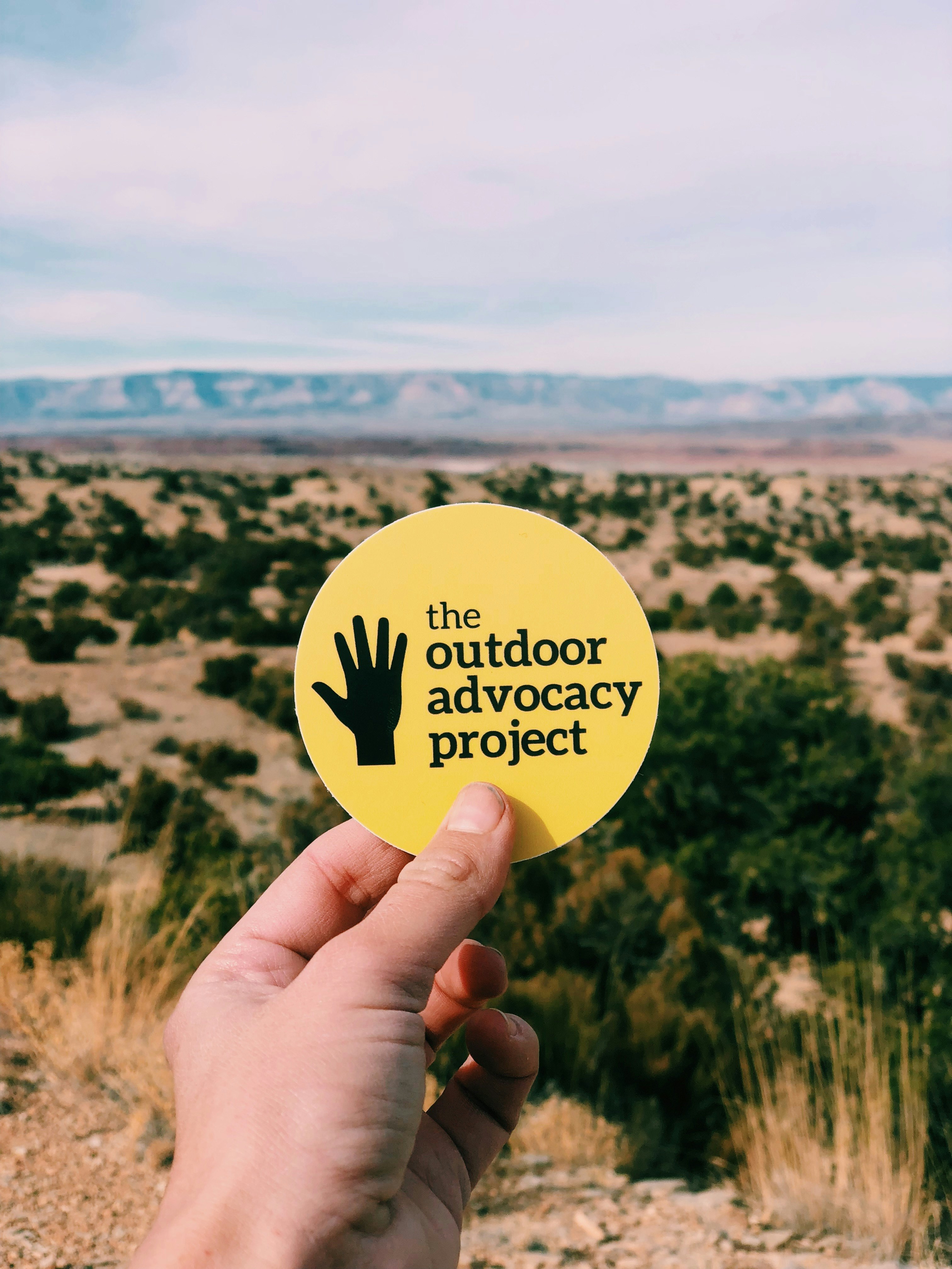 A white hand holds up a yellow Outdoor Advocacy sticker with an illustration of a black hand next to the text in front of a outdoor desert landscape