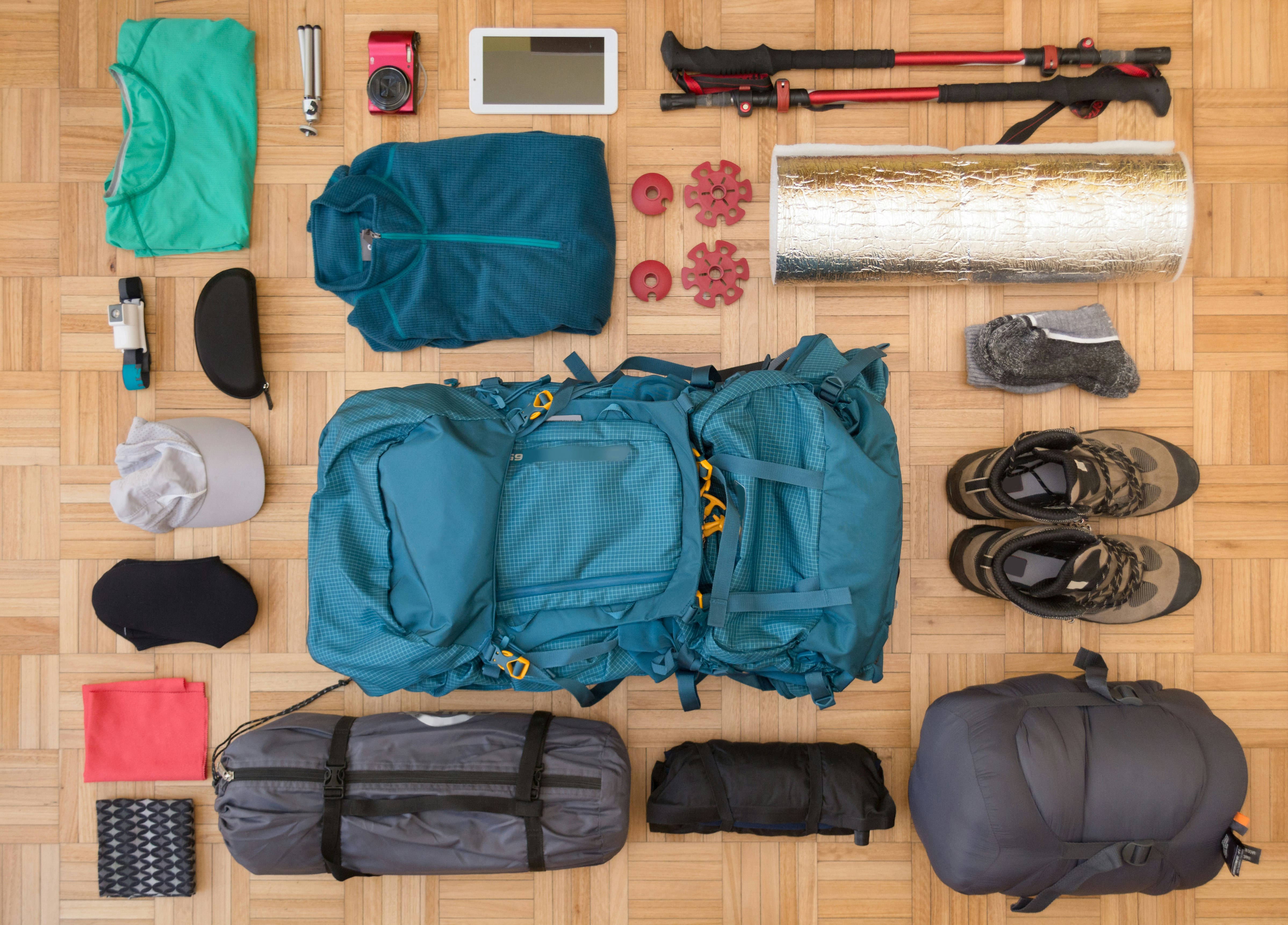 Individual items in a camping kit, including a backpack, bedroll, tent in a bag, boots, trekking poles, etc, are laid out on a parquet floor and shot from above, flatlay style