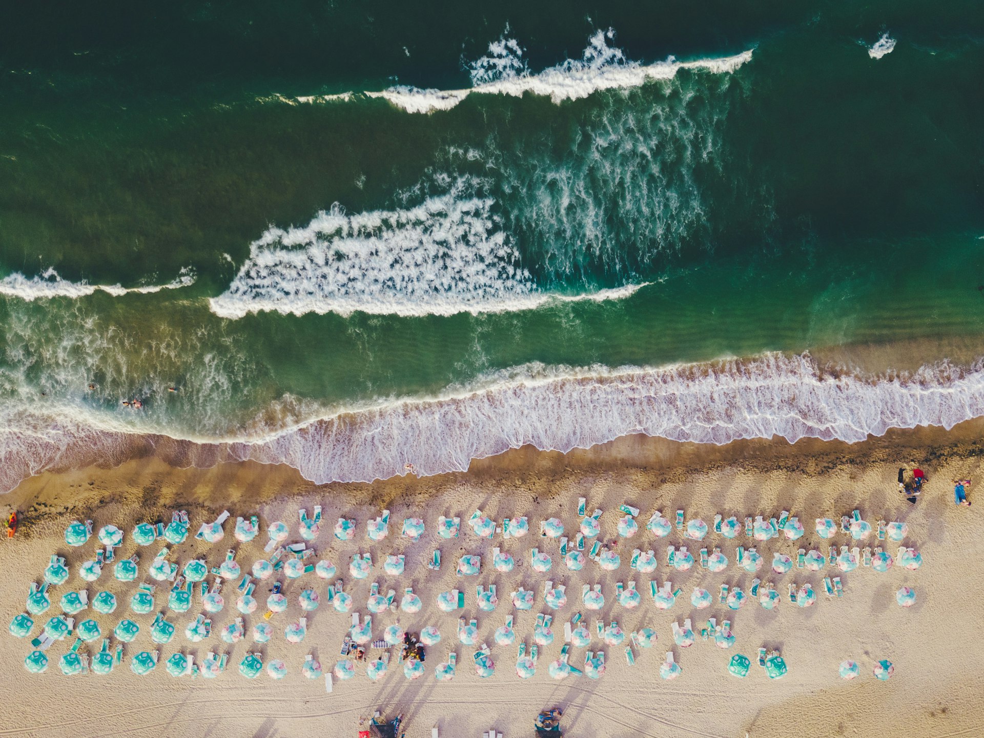 An aerial view shows bright umbrellas on the beach and a deep ocean in front. 