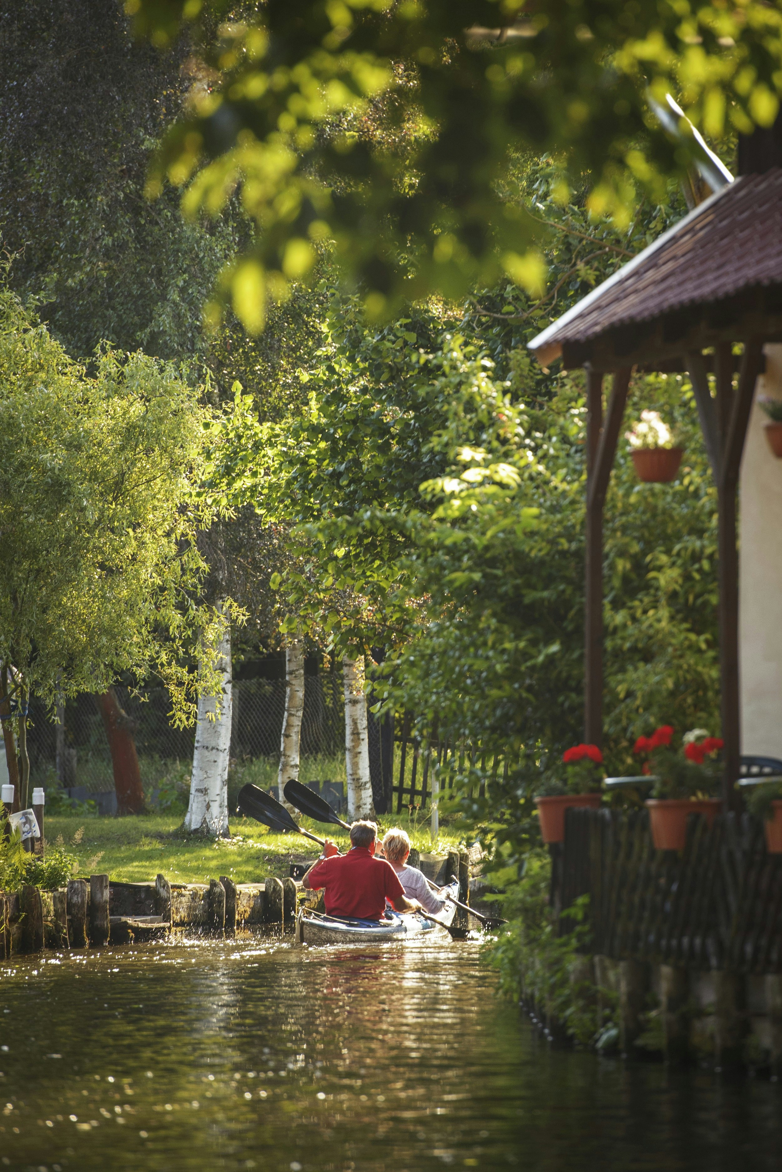 Two people kayak down a narrow waterway that cuts through grassy meadows dotted with trees and past houses perched on its edge.