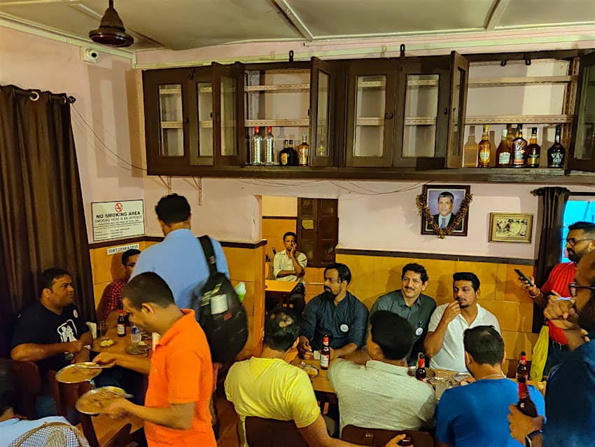 panjim-tavern-trail-goa.jpg Patrons gather in the white and ochre interior of Panjim Tavern, where brown wood cabinets holding bottles of liquor hang high up on the wall