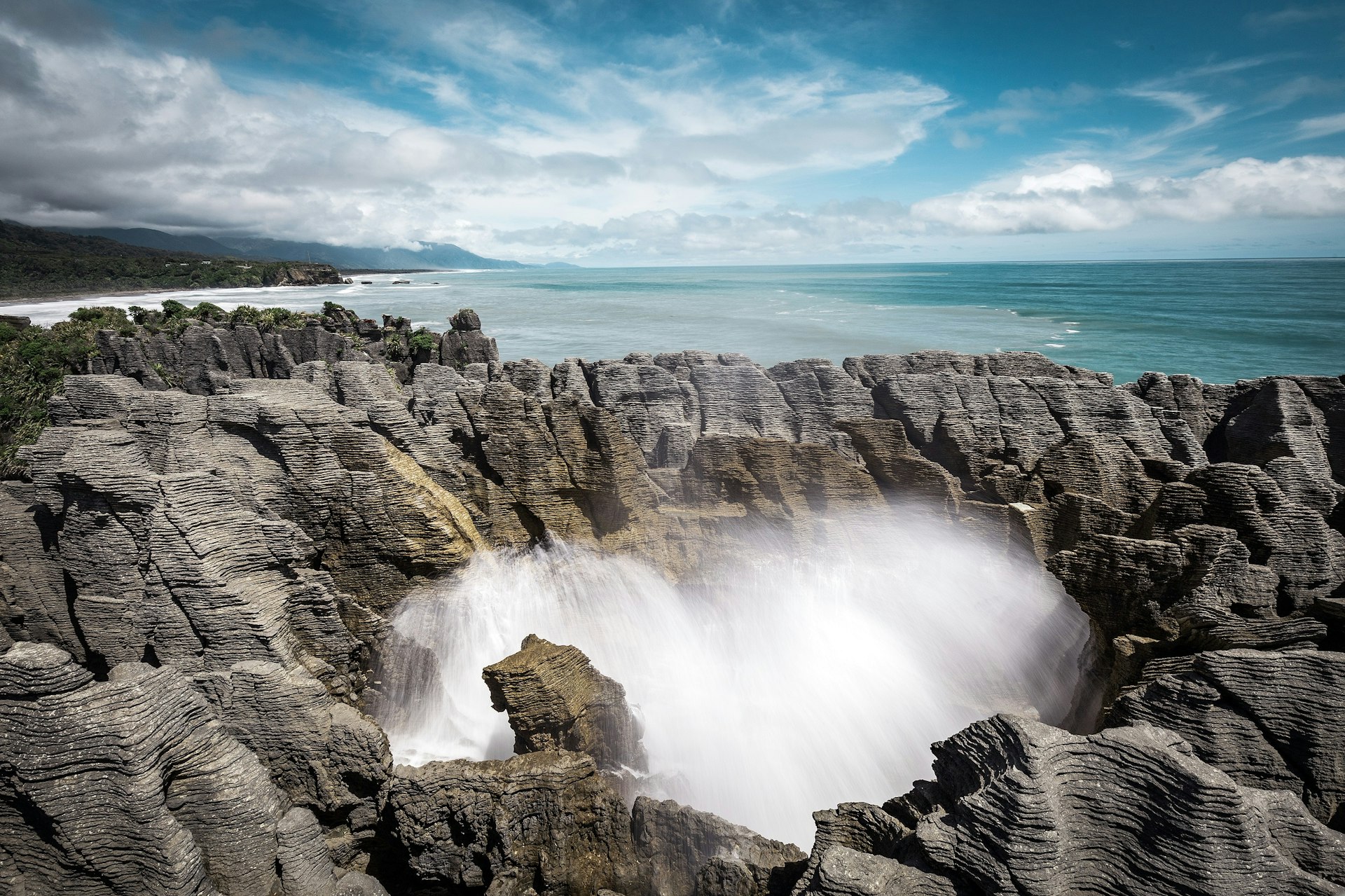 Formations of grey rock, seemingly made of thin sheets of horizontal 'pancakes' rise up into the sky; in the middle of the formation is a blow hole and water is shooting up. In the back ground is the Tasman Sea.