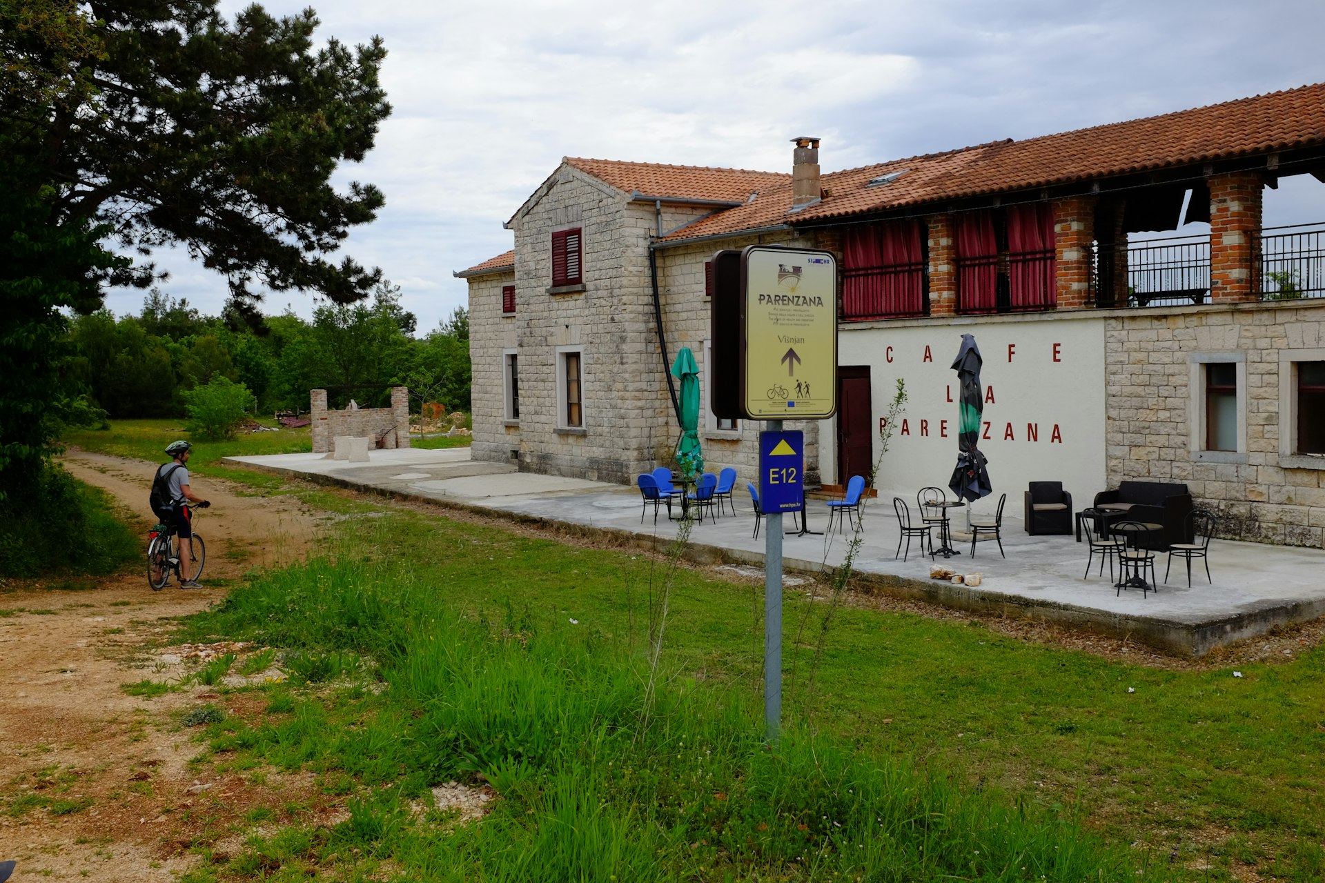 A cyclist stops on a dirt track of the Parenzana trail to glance at a former train station and siding that has been converted into a chic cafe. In the foreground is a sign indicating the track is the Parenzana trail for cyclists and trekkers