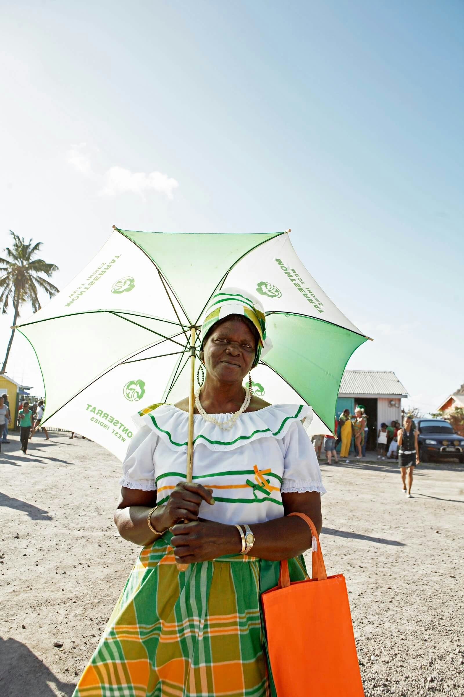 A woman dressed in orange, white and green holding a green and white umbrella on the beach on a sunny day