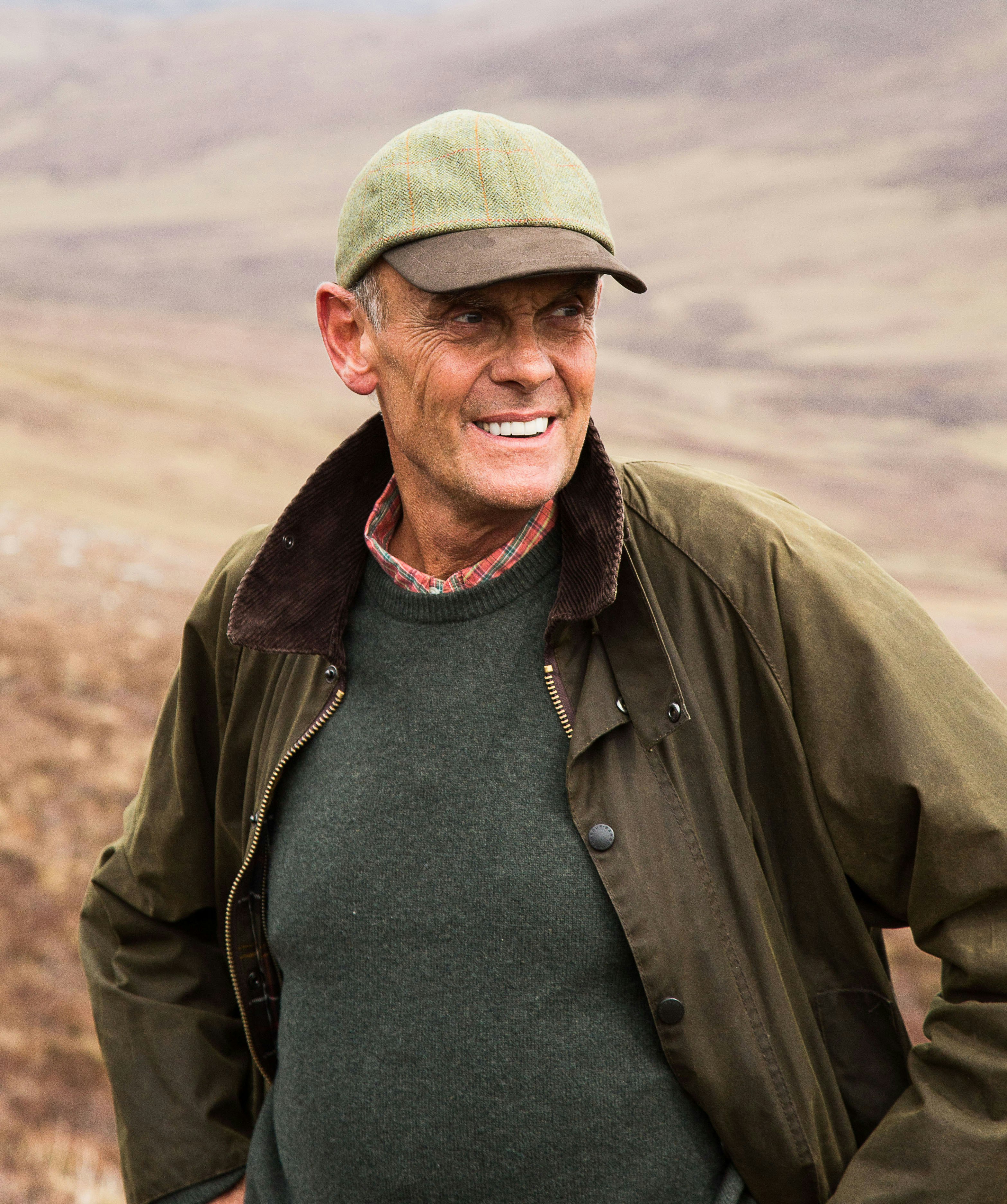 Paul Lister stands smiling; he's wearing a tweed baseball-style hat, with a red checkered shirt beneath a sweater and waterproof jacket.