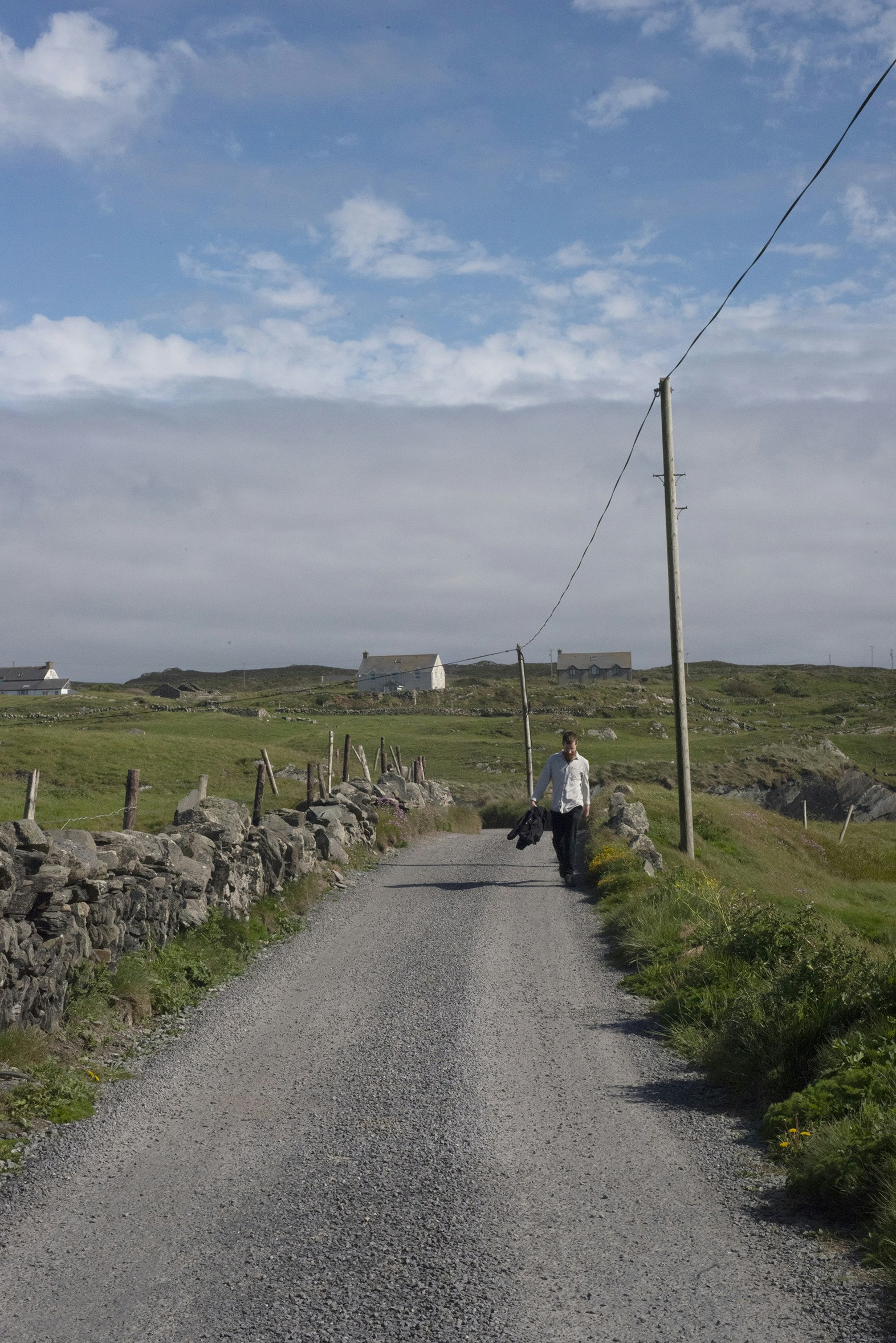 A man walks down a narrow rural road carrying his jacket. There are stone walls on either side and green fields all around.