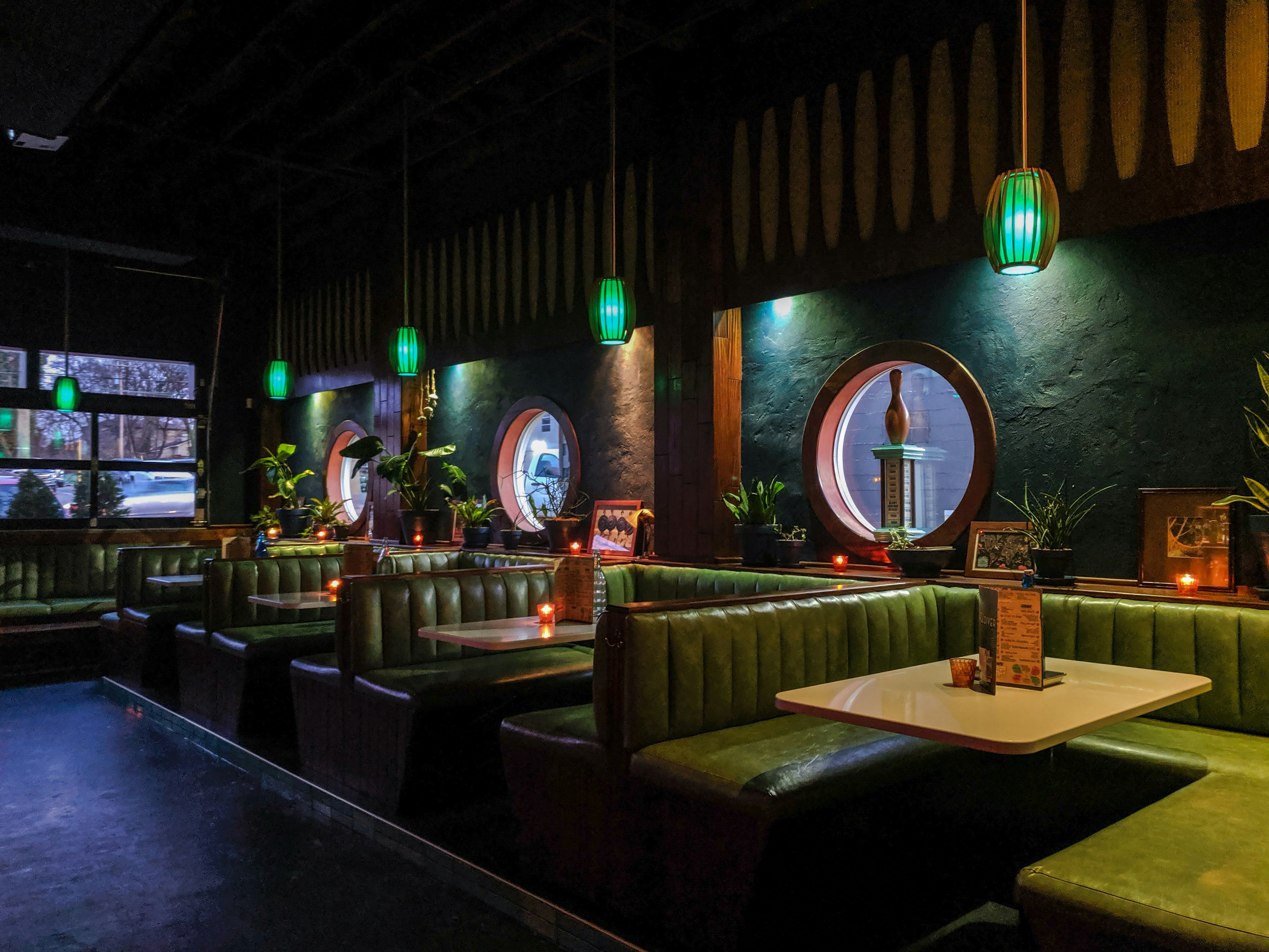 The dark, emerald green-tinted interior of Pearl Diver in Nashville, Tennessee features midcentury green naugahyde U-shaped booths against a hunter green plaster wall punctuated by porthole windows filled with bowling trophies and the like. Over the booths are hanging lamps in bottle-green glass shades. Live plants sit on a deep sill by the windows 