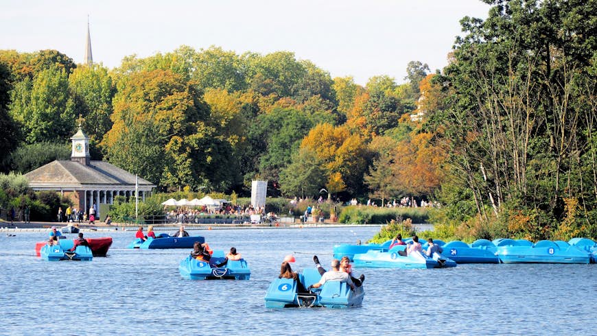 Five blue pedal boats are out on a lake on a summer's day. In the distance, tables are full of diners outside a cafe.