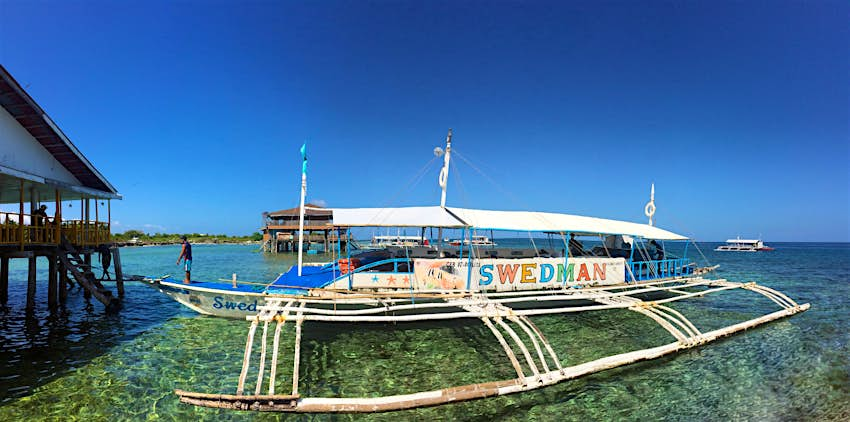 A worn white catamaran diveboat in the Philippines sits in shallow water dappled with light. In large rainbow letters on the side the name of the boat reads Swedman.