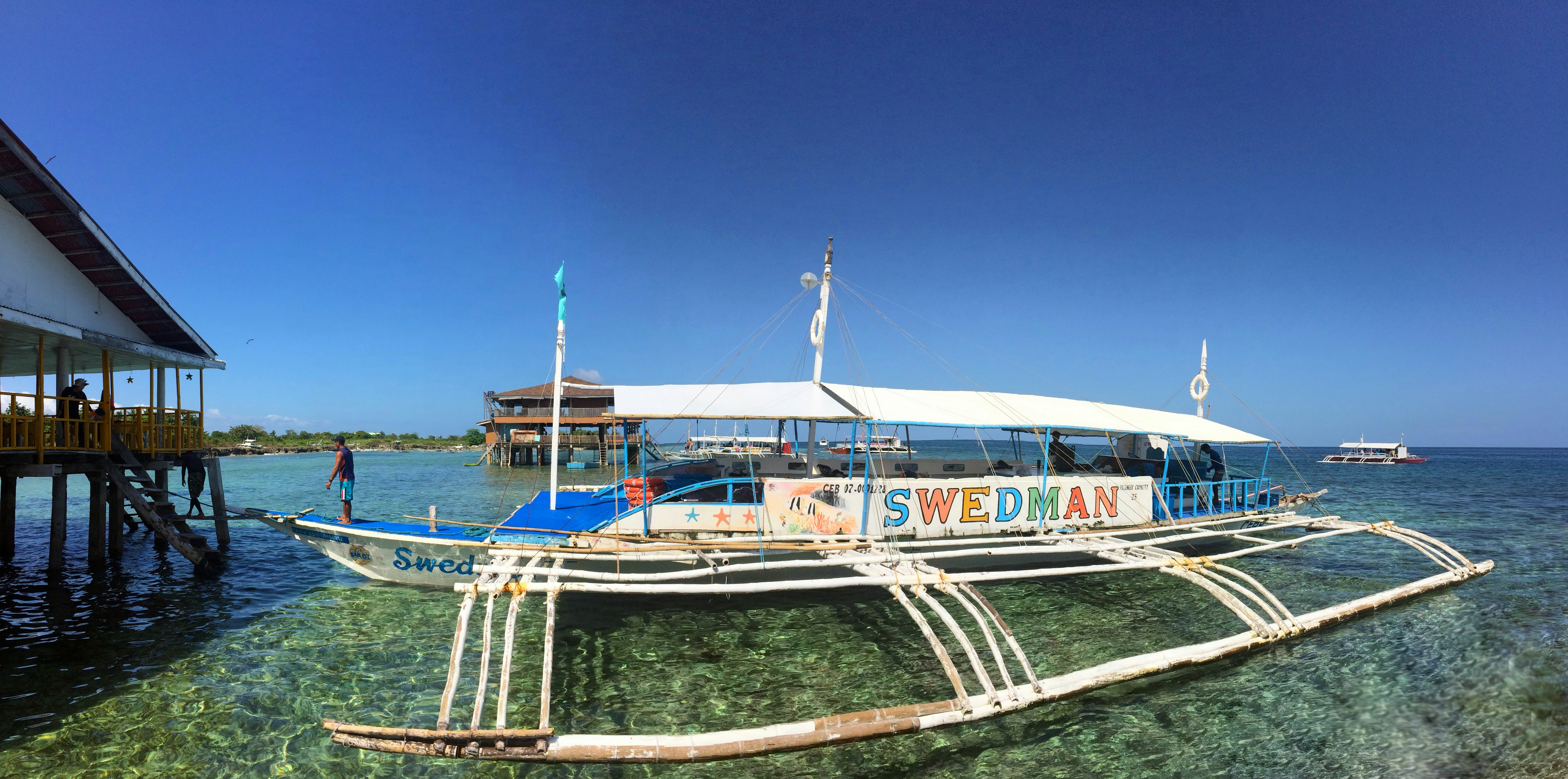 A worn white catamaran diveboat in the Philippines sits in shallow water dappled with light. In large rainbow letters on the side the name of the boat reads Swedman.