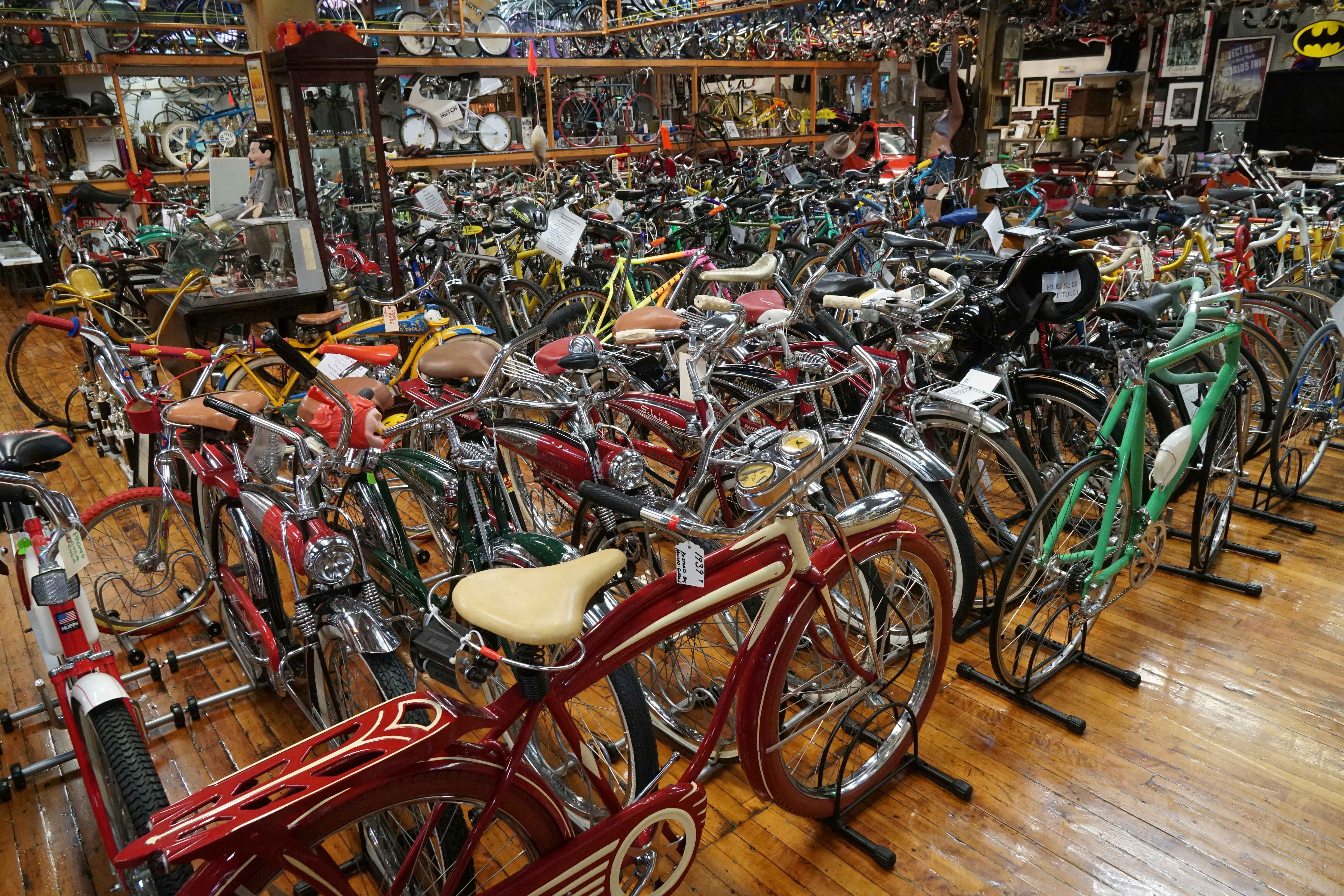 A room with glossy old hardwood floors is crammed with an inconceivable number of bicycles in all colors, shapes and sizes. In the foreground is a red bicycle with a cream seat and a teal one with a narrow, modern frame, but all you can see of the others are a jumble of seats, handlebars, and chrome gleam