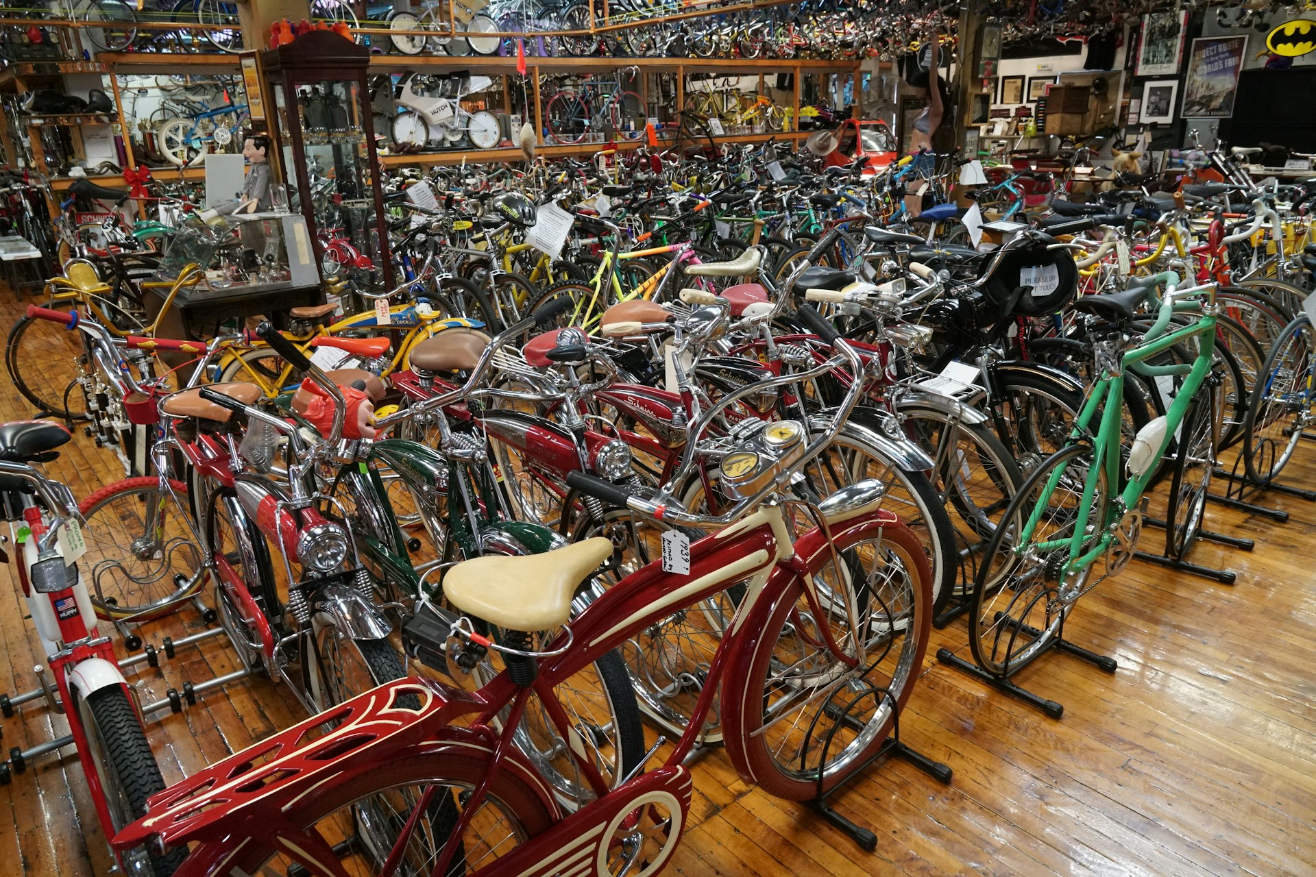 A room with glossy old hardwood floors is crammed with an inconceivable number of bicycles in all colors, shapes and sizes. In the foreground is a red bicycle with a cream seat and a teal one with a narrow, modern frame, but all you can see of the others are a jumble of seats, handlebars, and chrome gleam