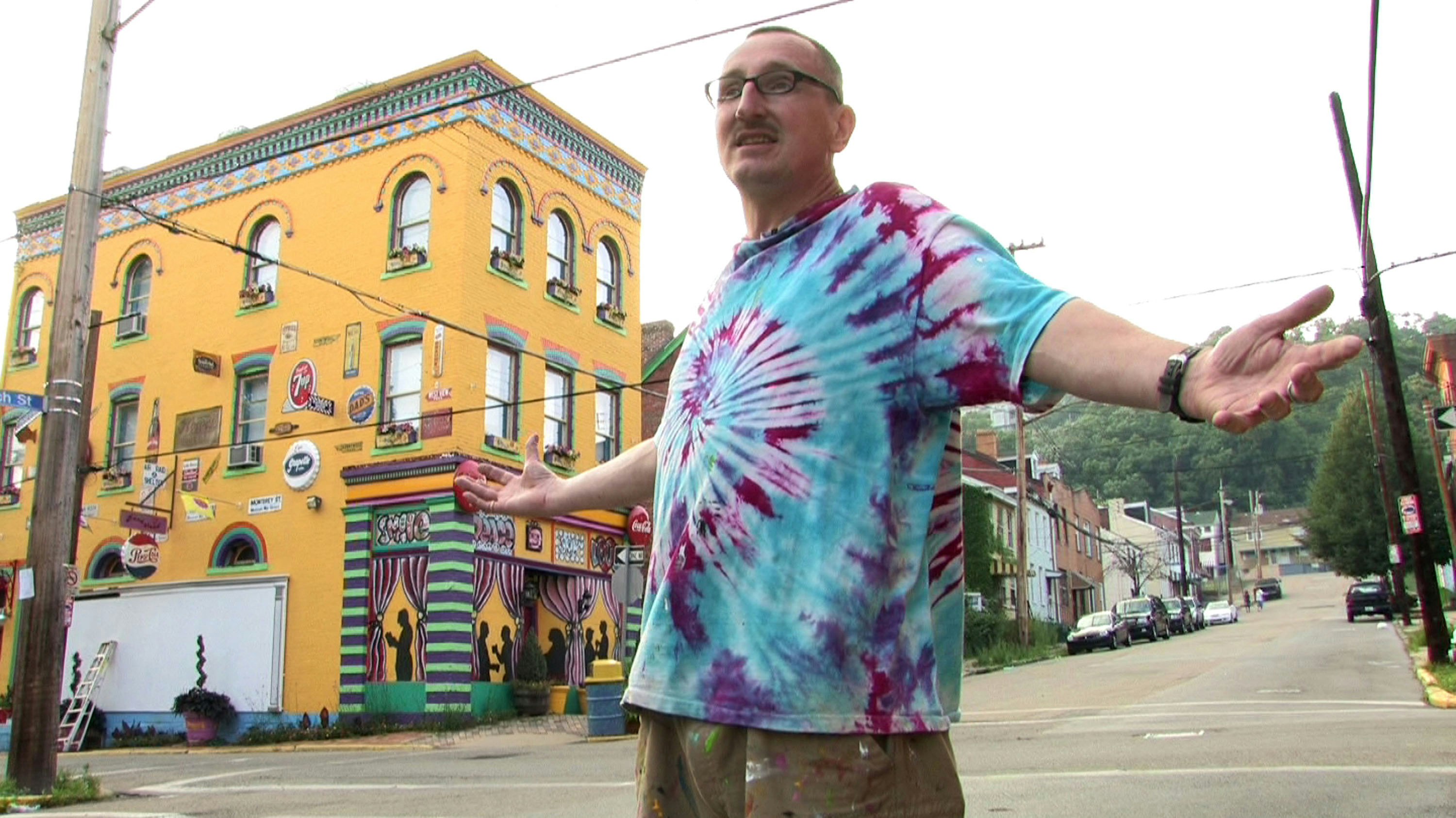 Street artist Randy Gilson stands in front of the yellow brick Italianate facade of Randyland, with his arms spread in a blue, red, and purple tie-dyed t-shirt
