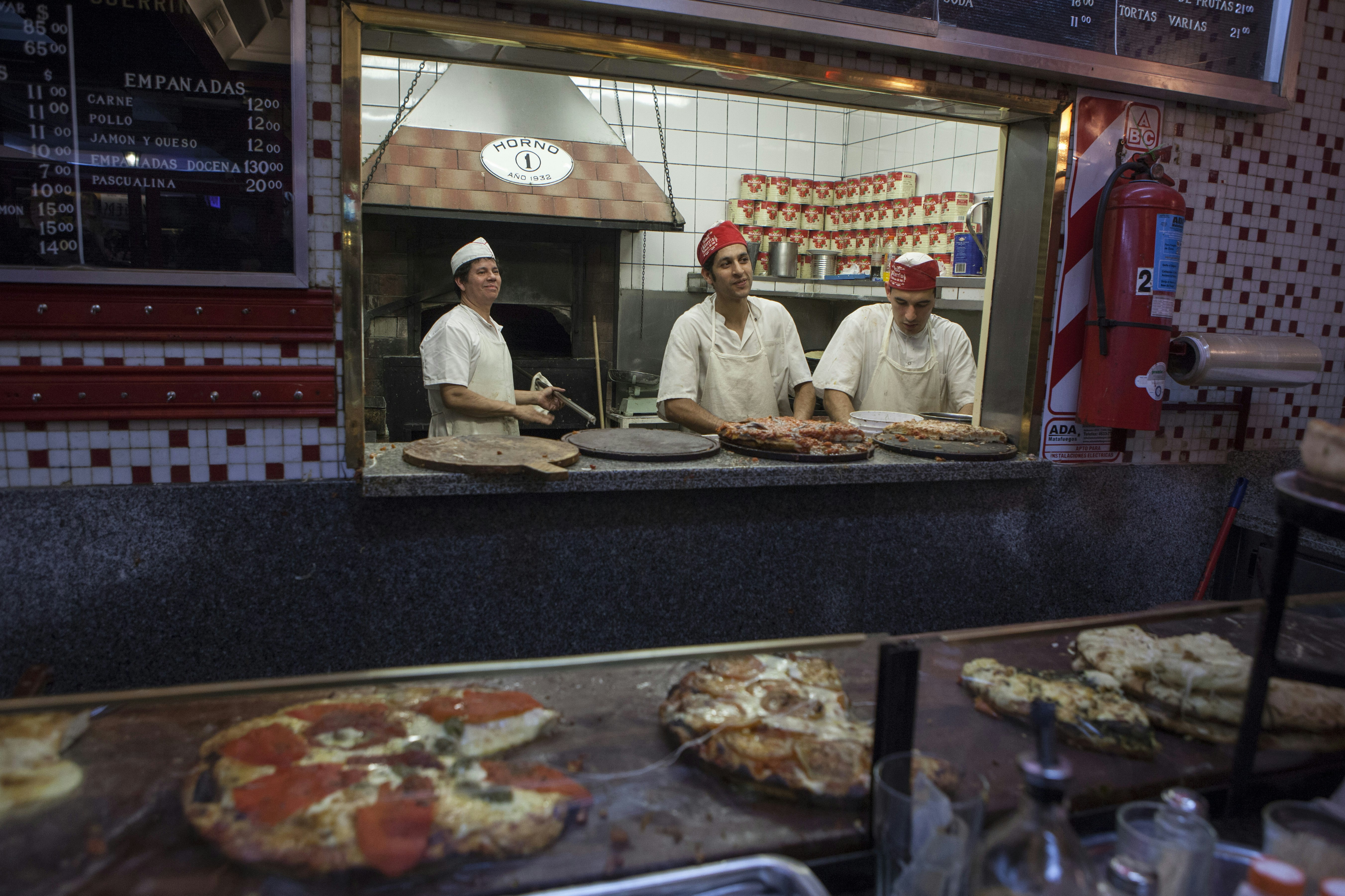 Three pizza chefs and a pizza oven can be seen through a hatch in the wall at Pizzería Güerrín. There are several pizzas laid out on a counter in the foreground.