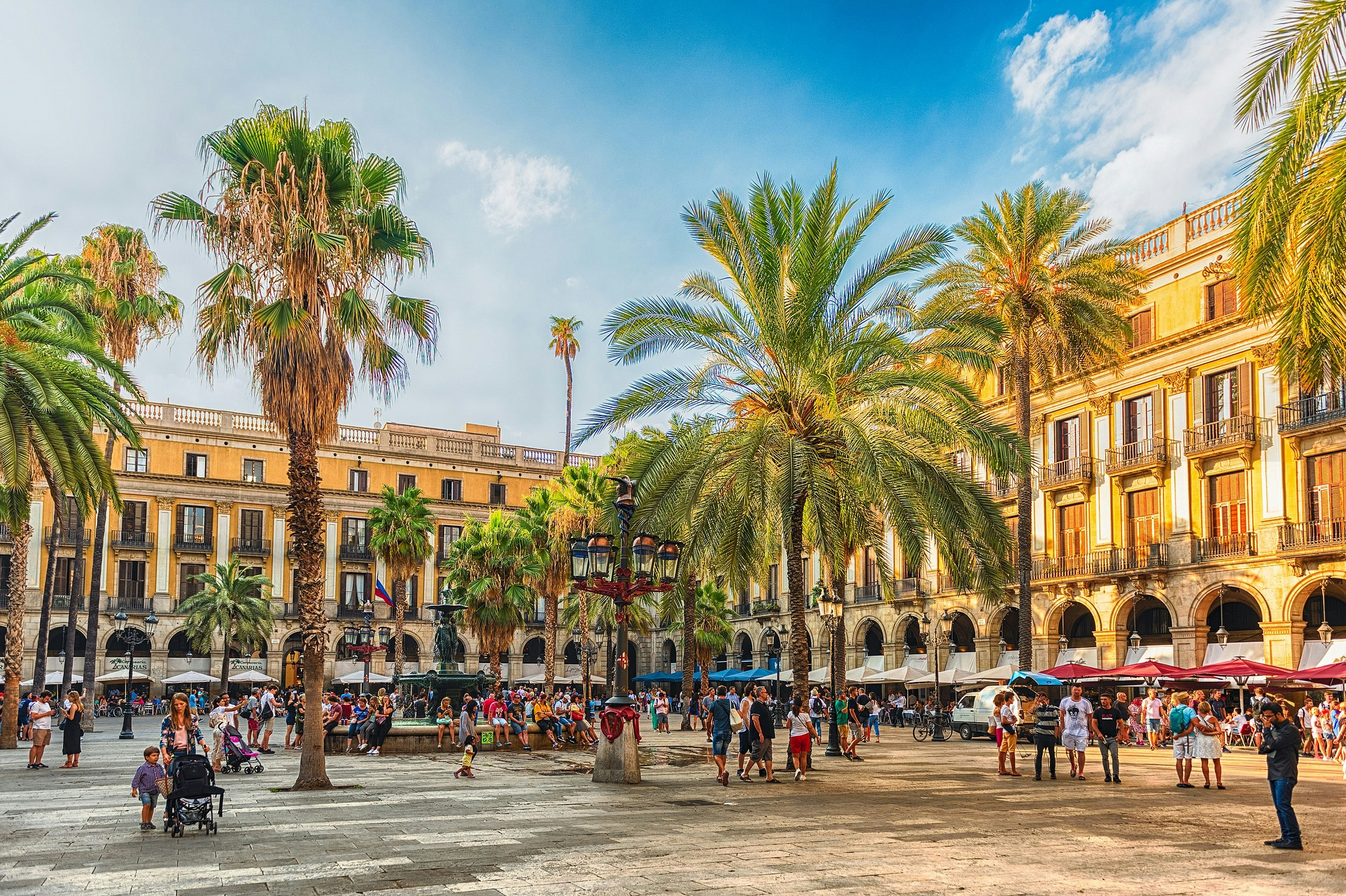 A large square with palm trees. The buildings lining the square are bathed in sunlight. Below each building are awnings and umbrellas. People sit at the restaurants there