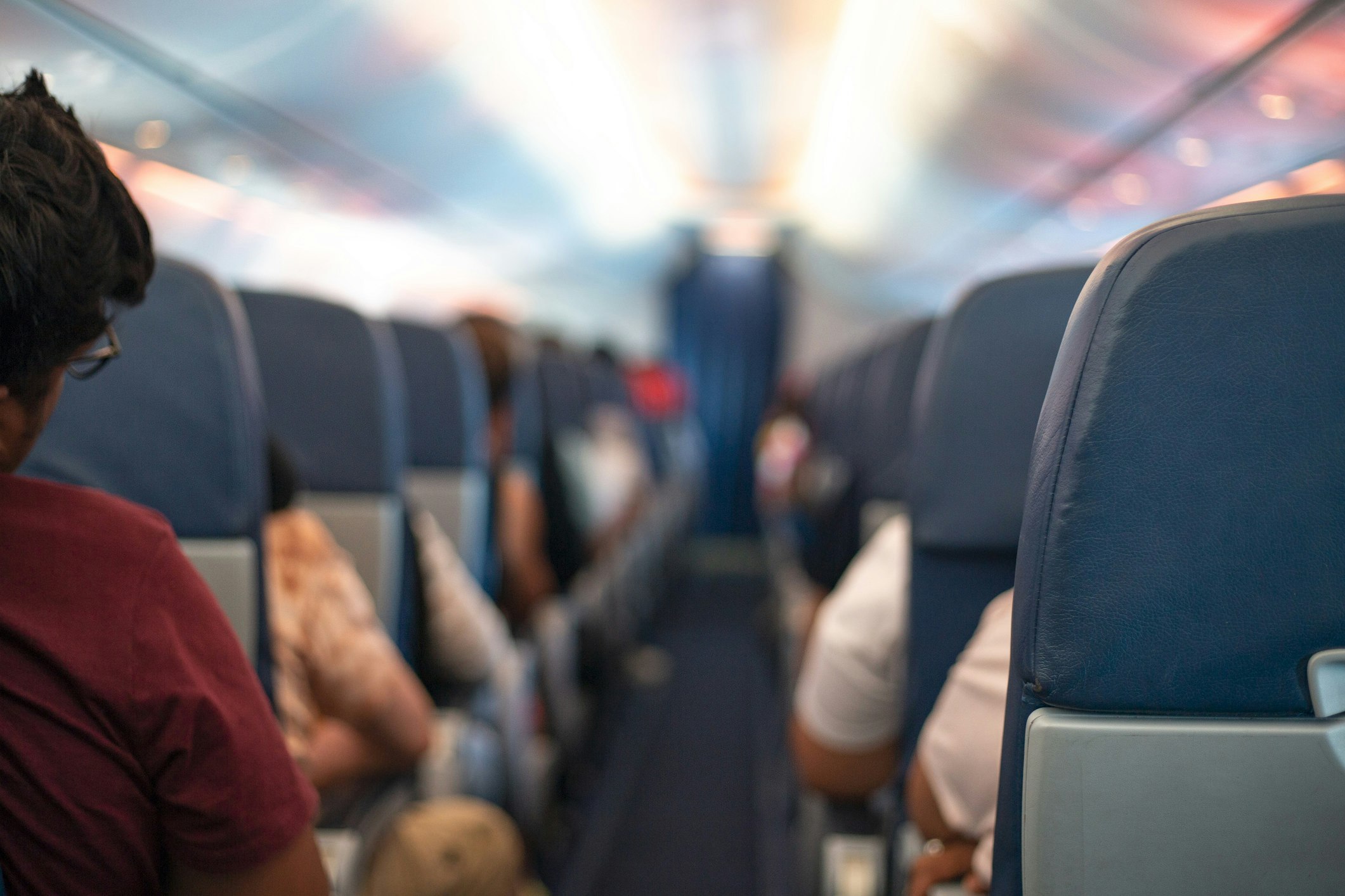 The interior of a plane from the aisle. The back of a woman's head and her right shoulder are partially visible from behind a seat. Other passengers are in shot but out of focus.