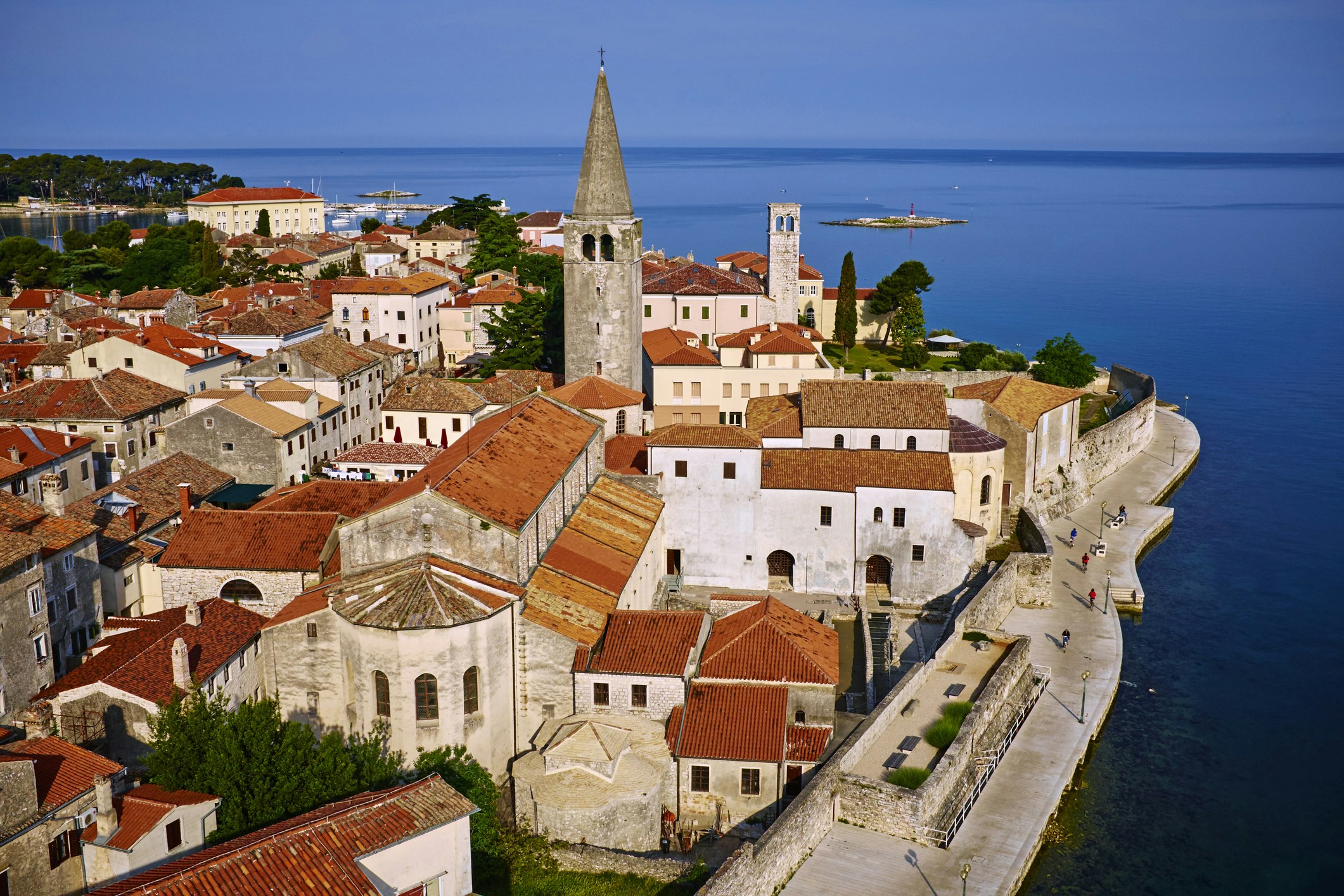 The red roofs and grey and white buildings of Porec, Croatia extend out into the deep blue sea, the same color as the sky on the horizon. In the forground you can clearly see the Euphrasian Basilica, its tall bell tower opposite the viewer