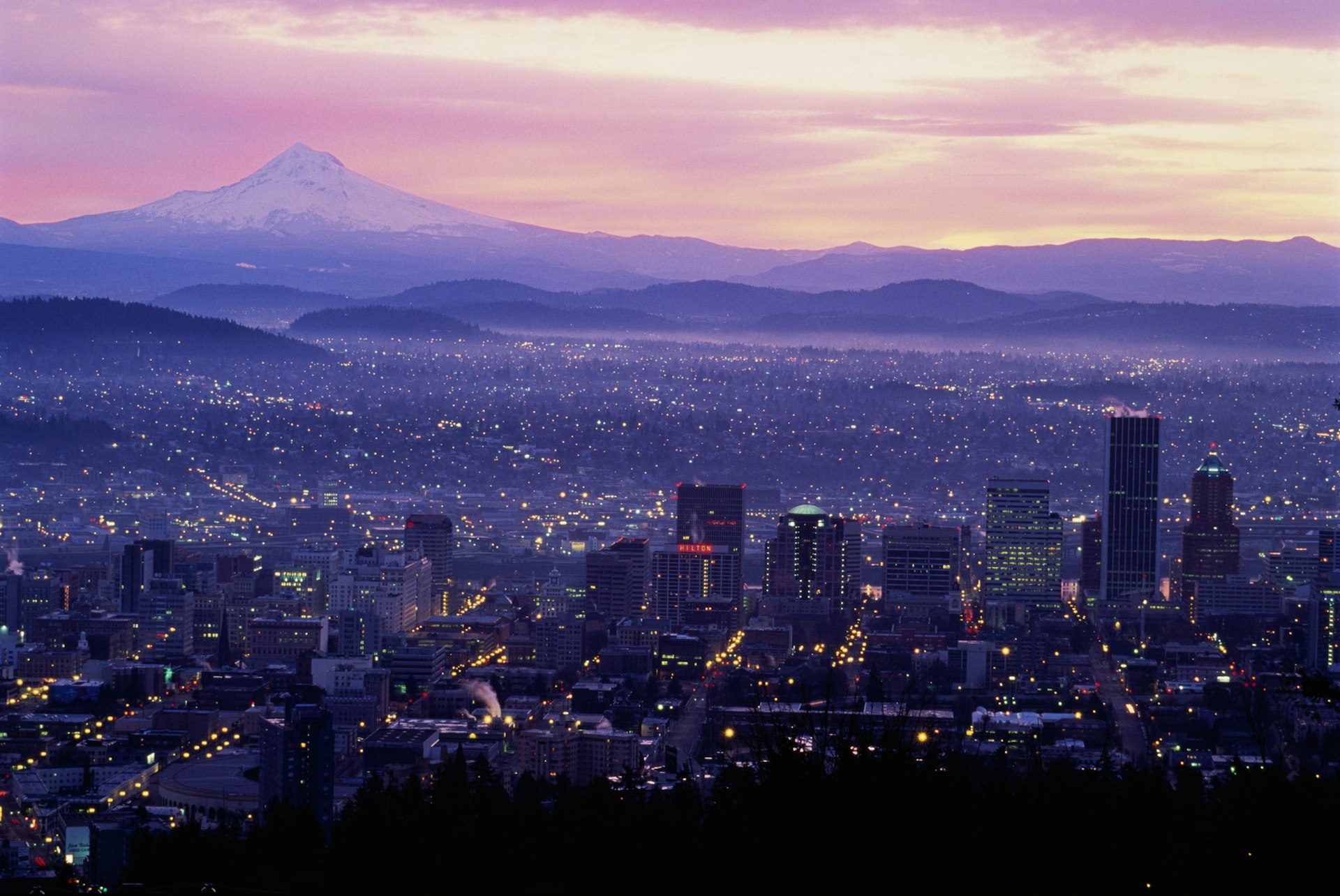 The sun rises over Portland, Oregon, washing the city's downtown skyscrapers and suburban sprawl in lavender and pink light as Mt. Hood rises over the landscape in the top left of the frame