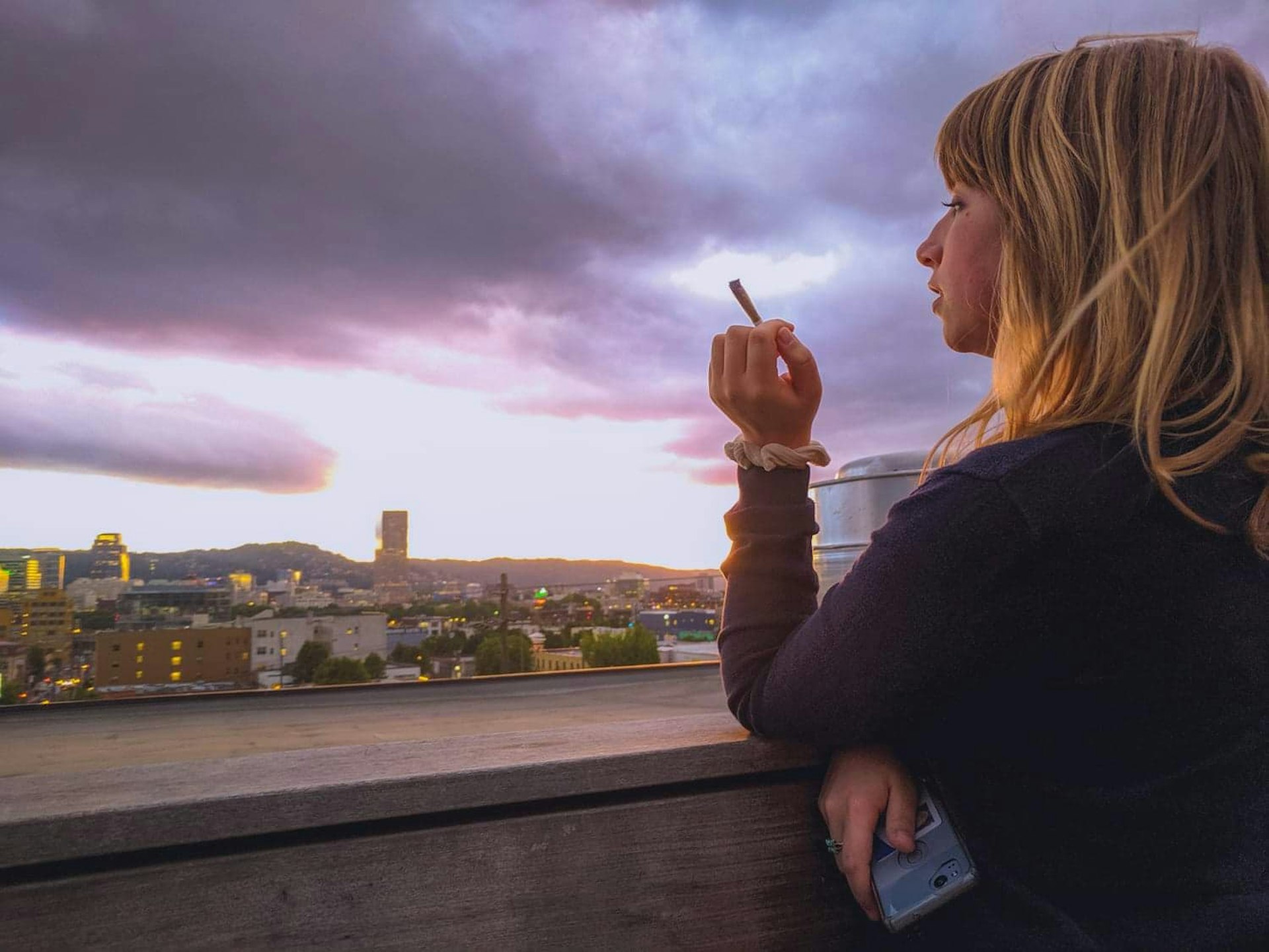 A young woman with red hair holds a joint in her left hand as she looks out over the Portland, Oregon skyline at dusk