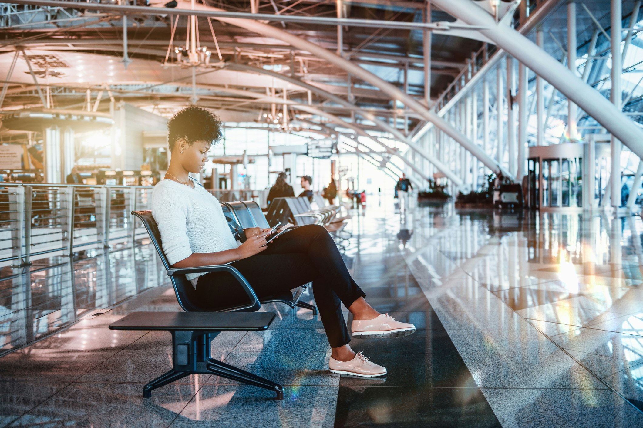 A black female tourist with a short natural hairdo, a white sweater, black slacks and pink slip-on shoes sits in the Porto airport in Portugal looking at her tablet.jpg