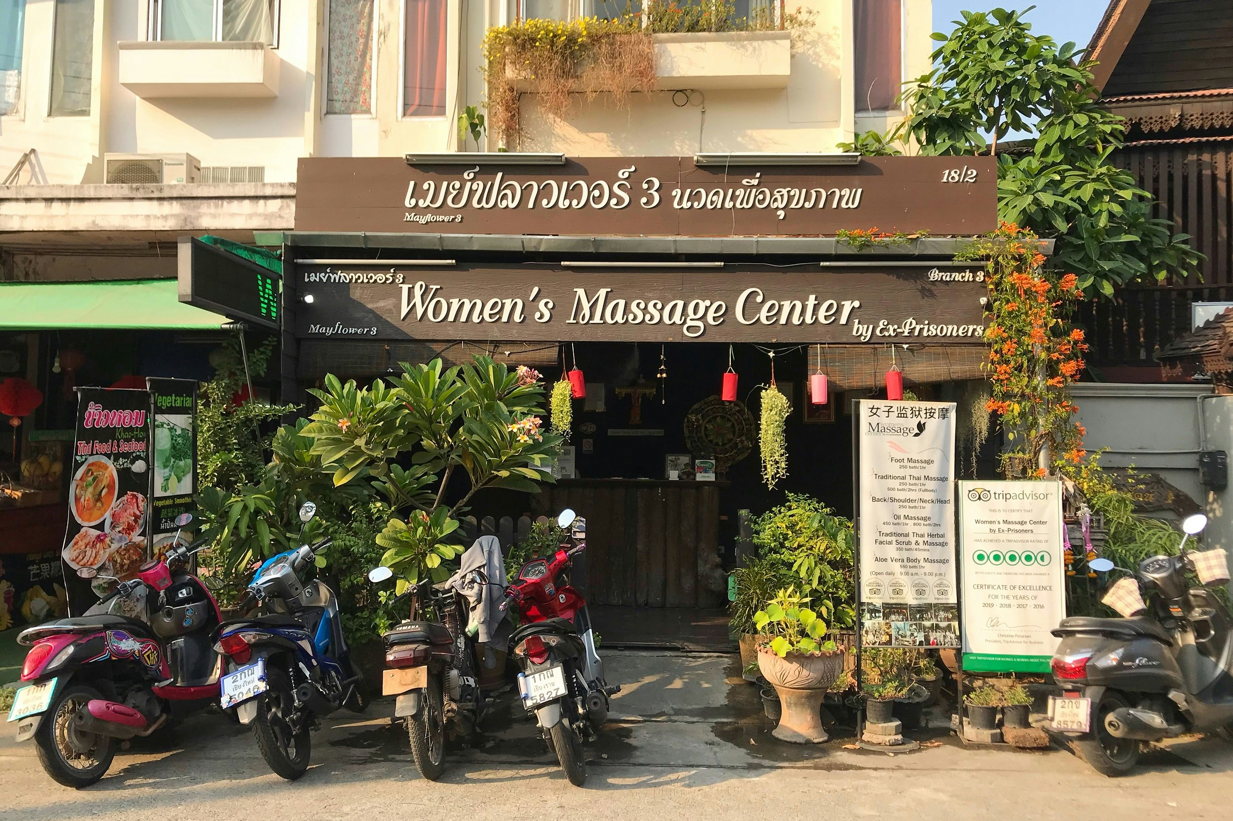 Front of a shop with a large sign in Thai and English that says "Women's Massage Center by ex-prisoners".  Five motorcycles are parked in front of the shop.