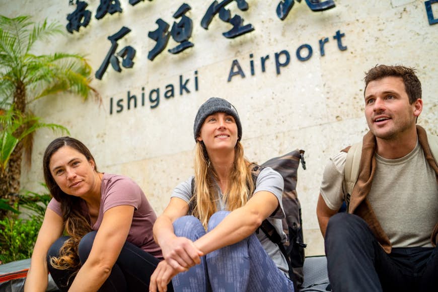 Två kvinnor och en man sitter utanför Ishigaki flygplats, kvinna i mitten bär pranas blå Izzie-mössa