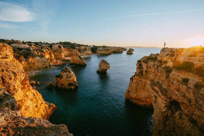 Portugal's rocky coastline at Praia da Marinha is bathed in warm sunlight; stacks of rock rise from a calm sea, and a figure is visible standing on a clifftop.