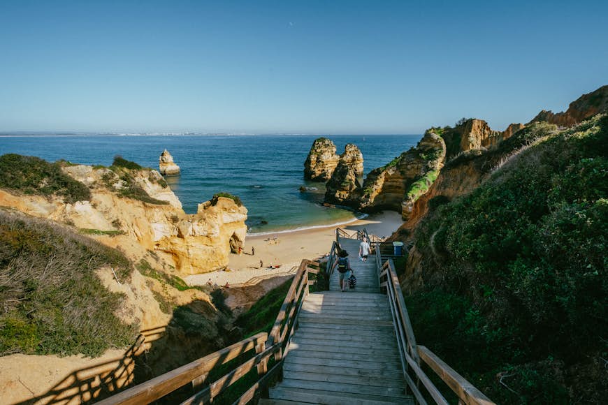 A few people walk down a set of wooden steps towards the sheltered sands of Praia do Camilo.
