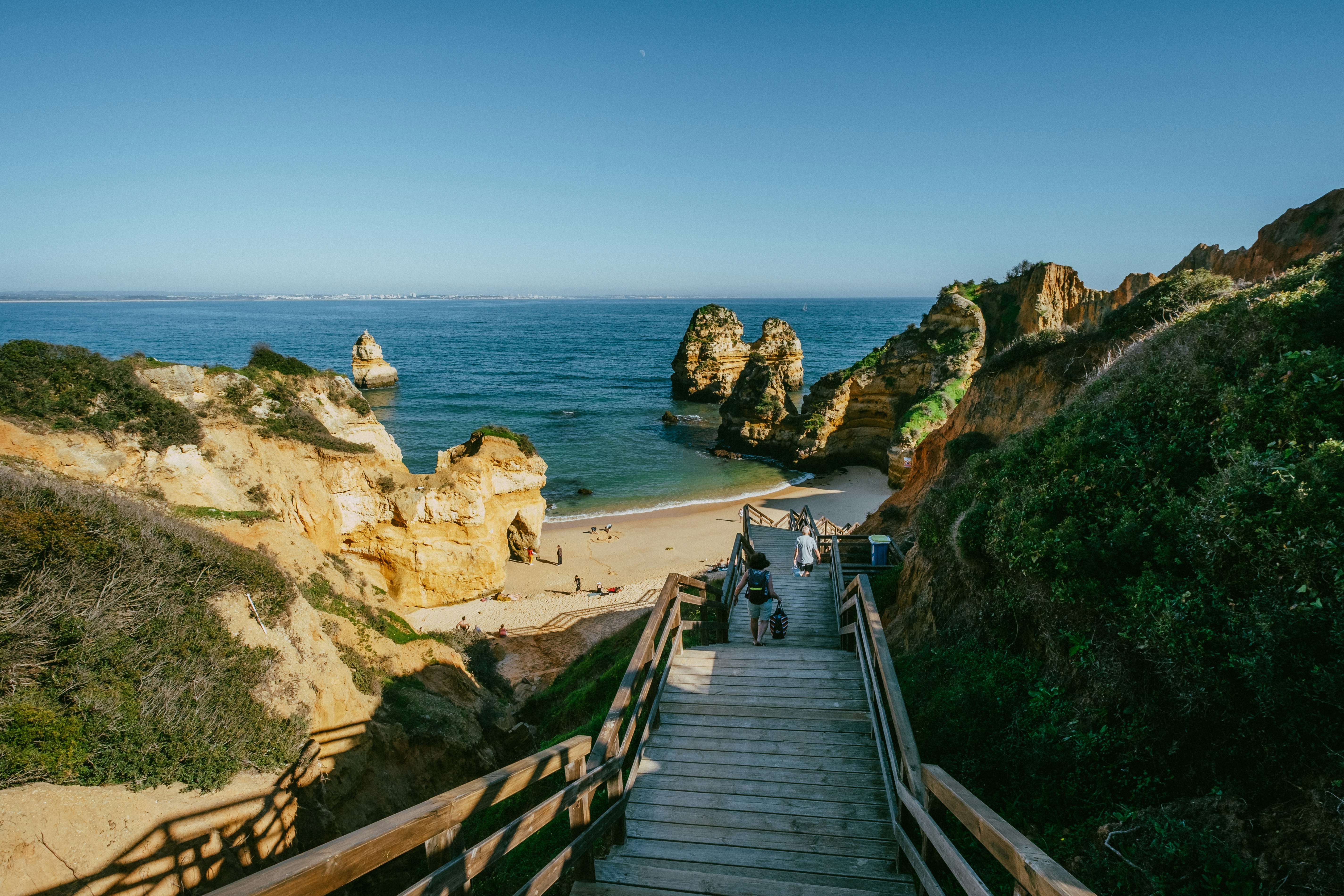 A few people walk down a set of wooden steps towards the sheltered sands of Praia do Camilo.