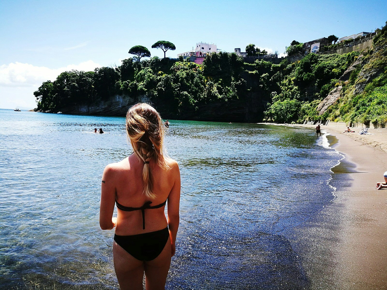 The rear view of a woman in a bikini facing the calm sea, with the beach arching out to her right; at the end of the beach are rocky cliffs topped by verdant forest.