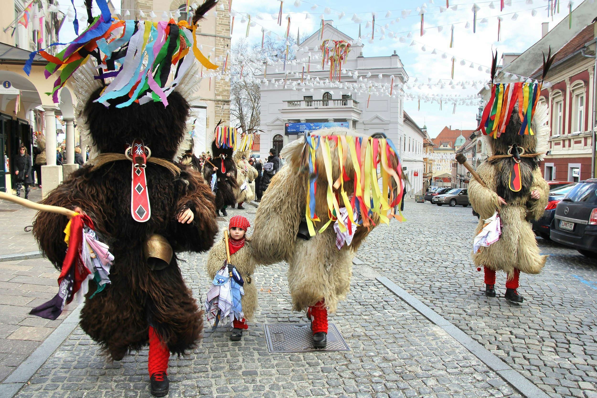 A procession of adults in large furry costumes with masks and headdresses makes its way down a cobbled street. A child wearing the furry costume and a red bandana walks between them