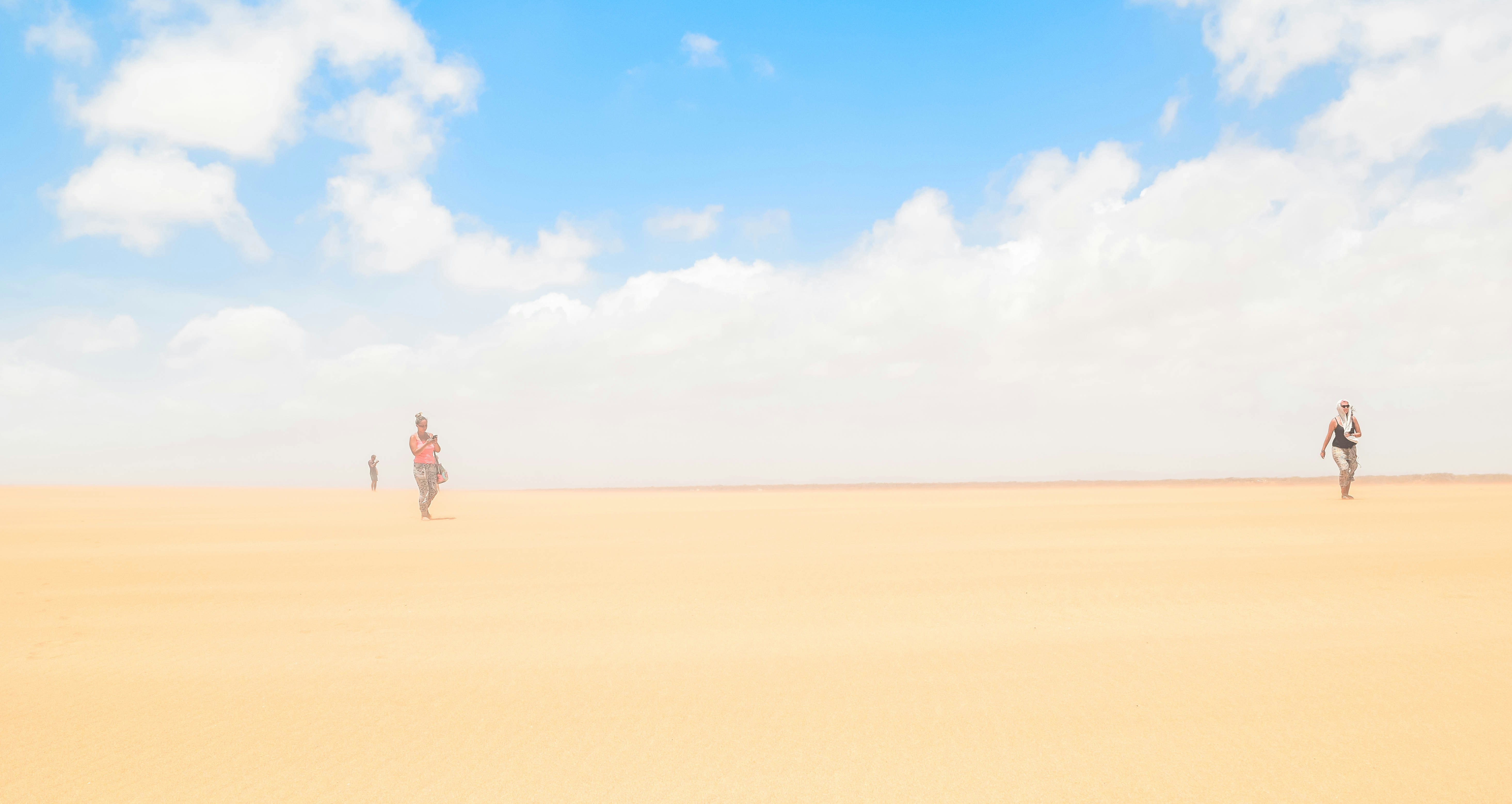 Three people, spread out across the image, walk across a vast expanse of straw-colored sand under a bright blue sky. The wind has whipped the sand in the air, and the figures are partially blurred.