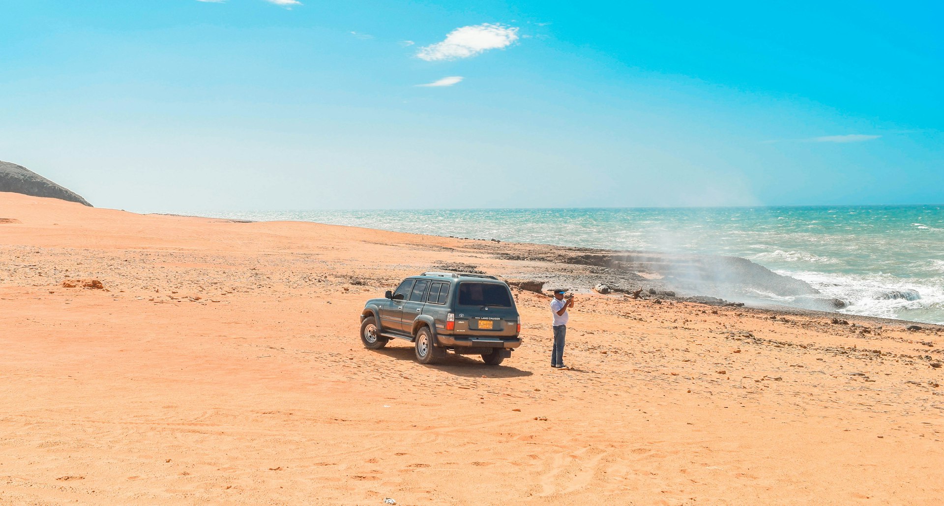 A 4WD vehicle is parked on sand on the Caribbean coastline; a guide is standing beside it, taking a photograph of the view out to sea.
