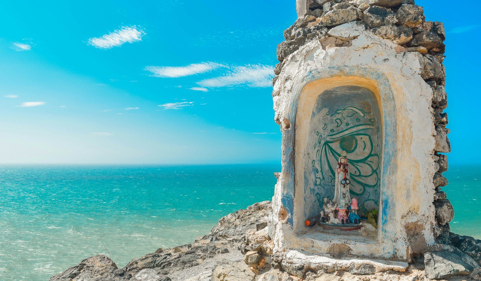 A small alter containing religious figures has been created out of rocks and stands at a viewpoint overlooking the dazzling azure Caribbean Sea.