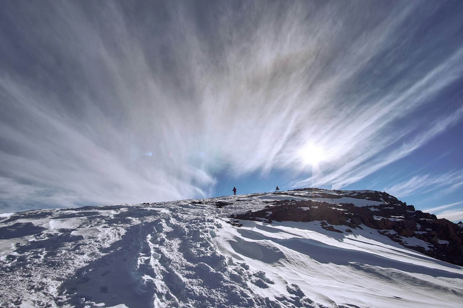 This image looks up a path in the snow towards a triangular cairn at the summit of Jebel Toubkal; a climber is seen approaching the top. The blue sky has wispy clouds.