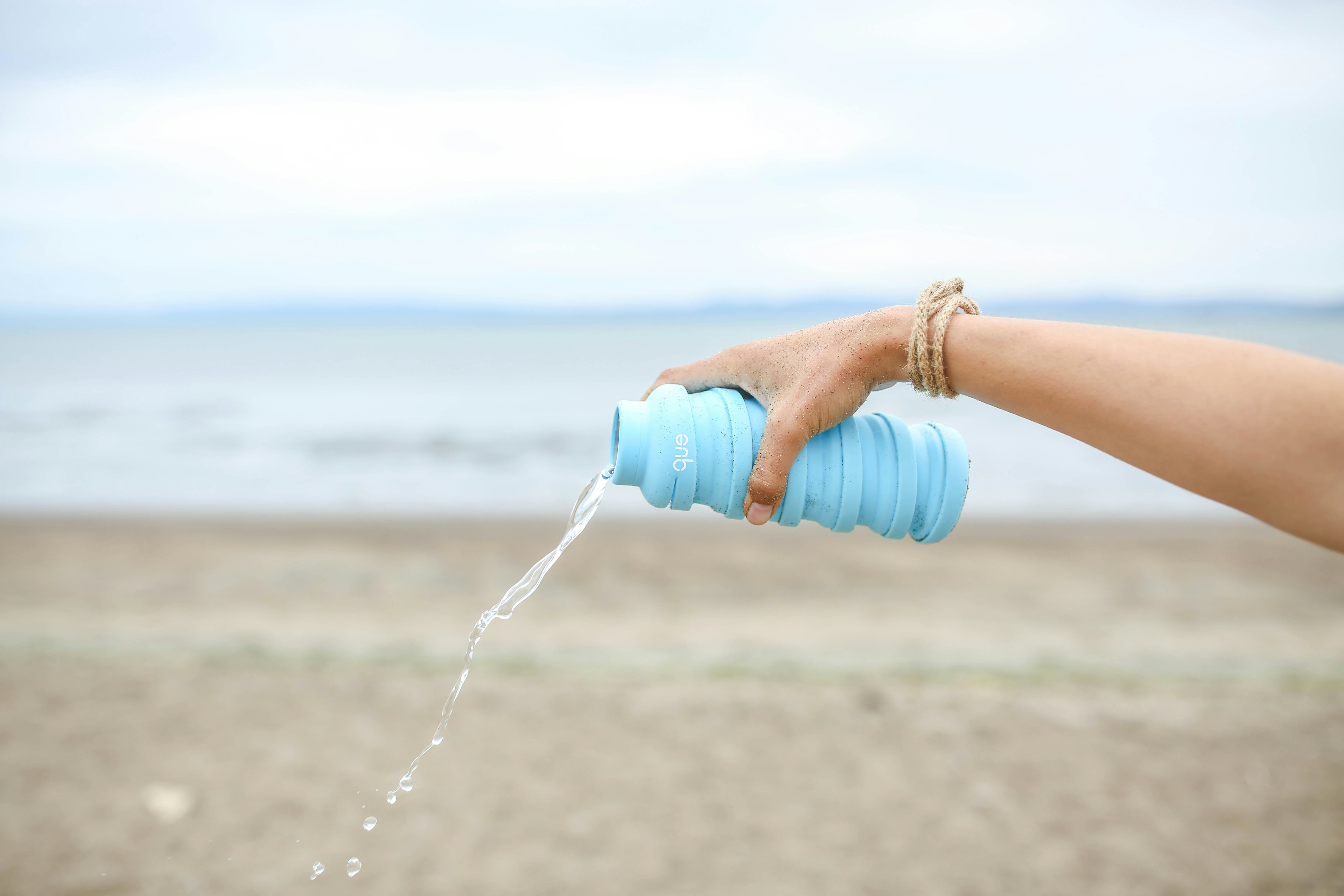 a hand holding a blue que bottle, pouring water out onto the beach