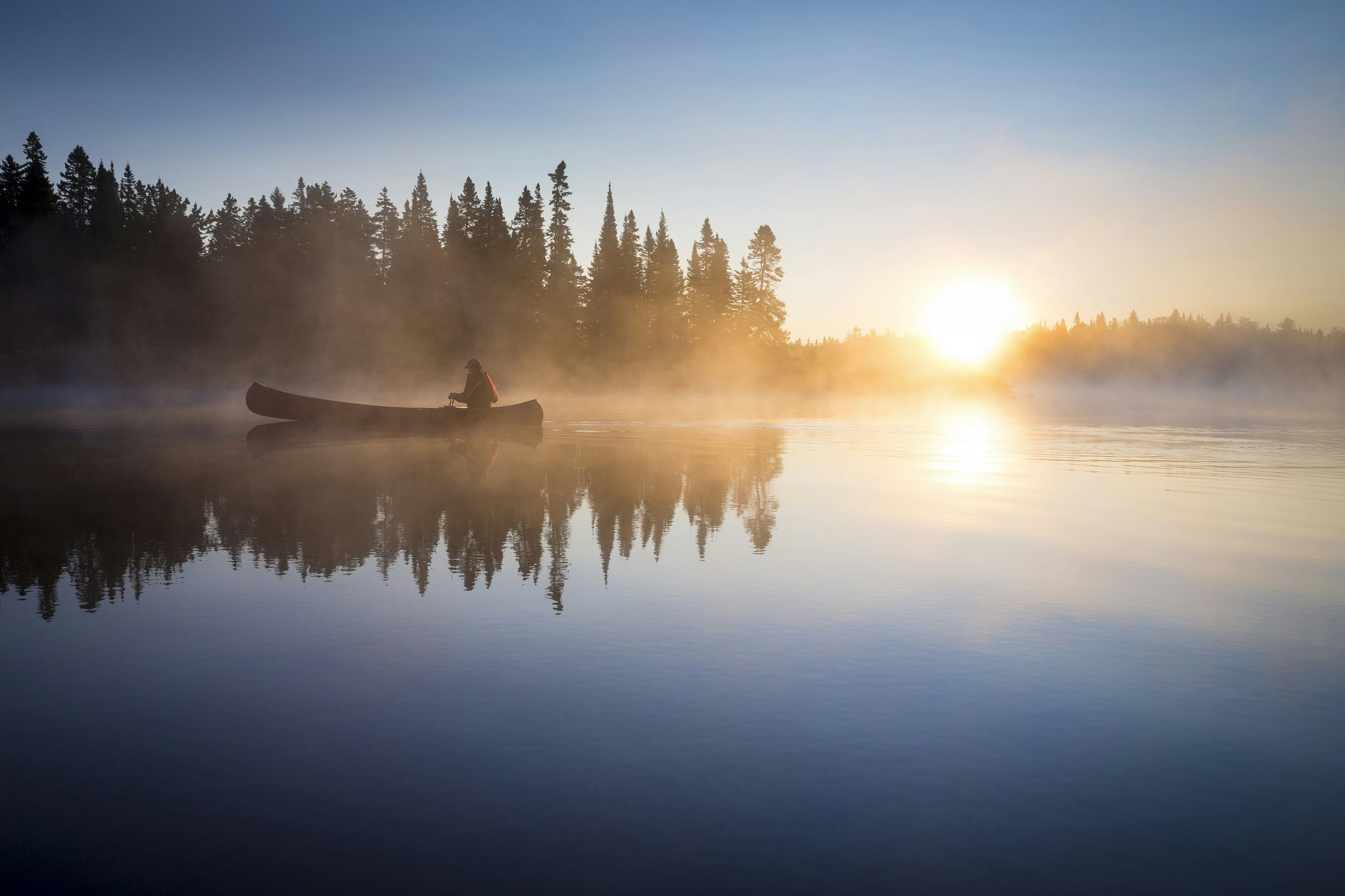 A single paddler canoes across a lake at sunrise; a light blanket of mist covers surface, with the silhouetted trees reflecting off the mirror-calm lake. The sun pierces the horizon, just above the treeline.