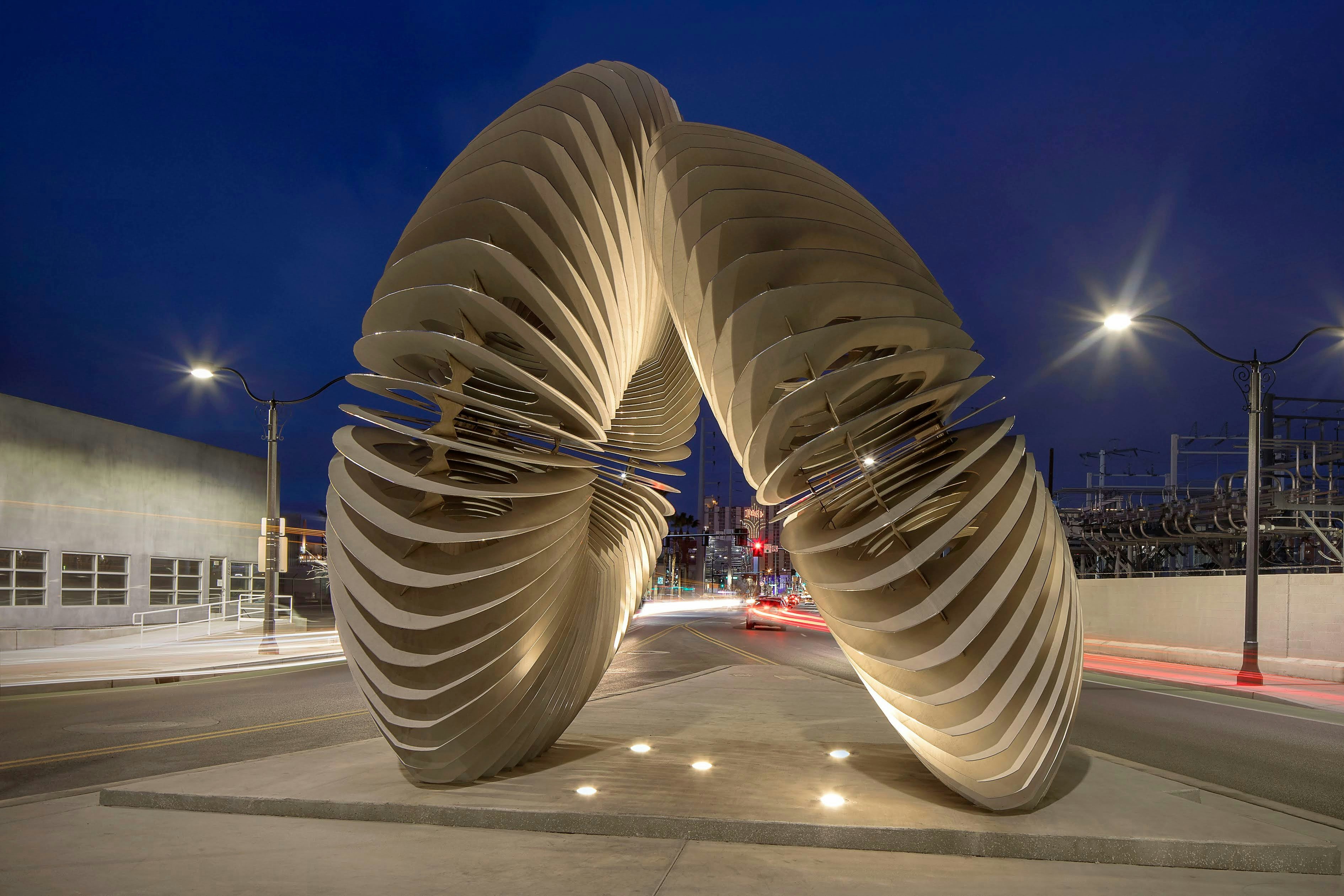 Luis Varela-Rico’s 'Radial Symmetry' sculpture at night; two large, circular forms, comprised of individual metal sheets, lean against one another on the sidewalk. 