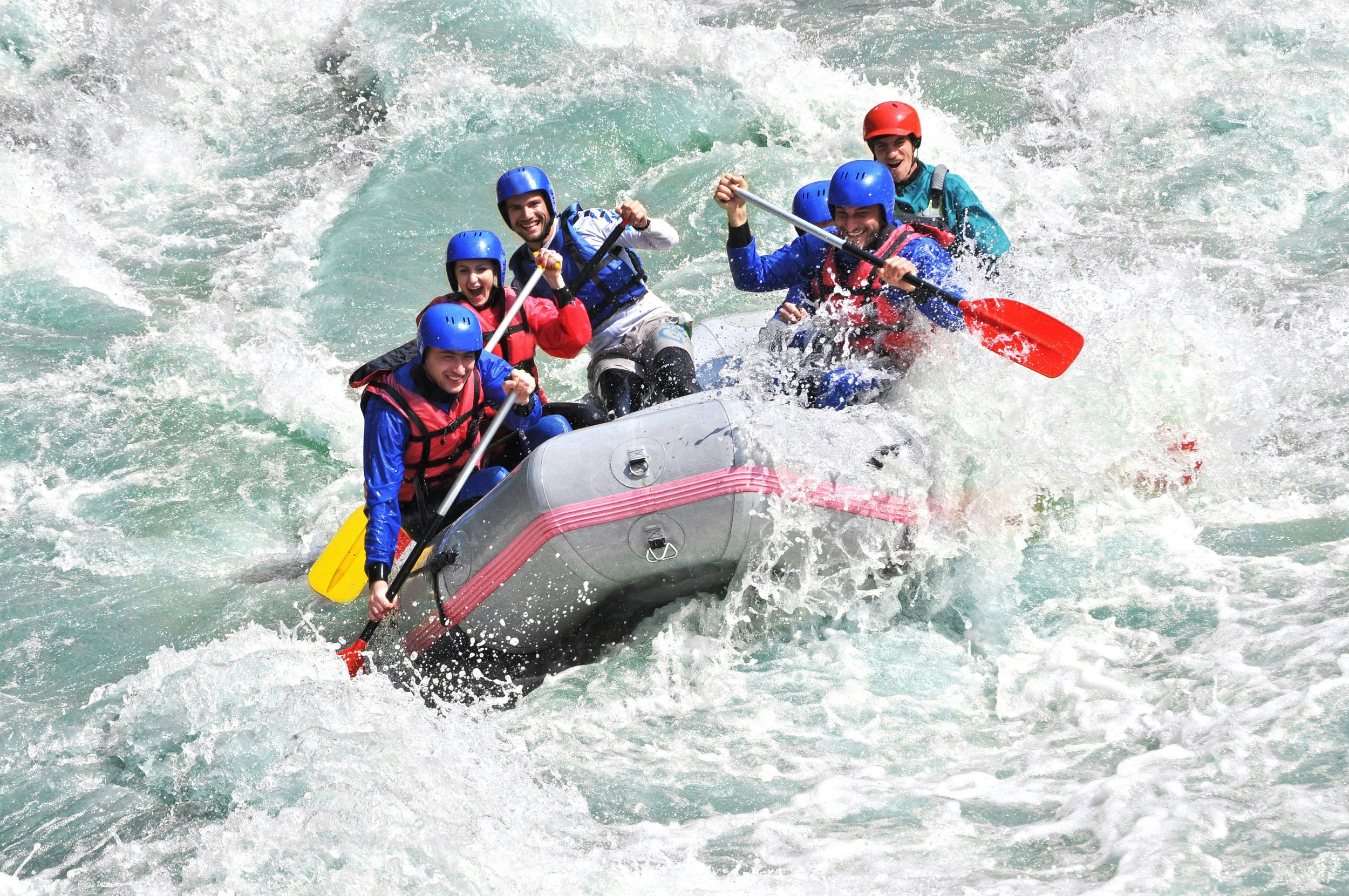 An inflatable raft with six smiling people in it crashes through whitewater; all are wearing blue helmets and red life jackets.