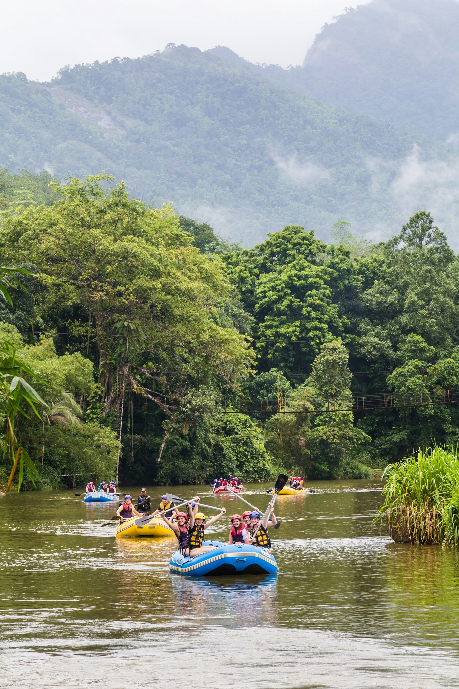 Several inflatable rafts, each holding six people, float along a serene section of river; the banks are lined with lush forest, as are the mountains in the background.