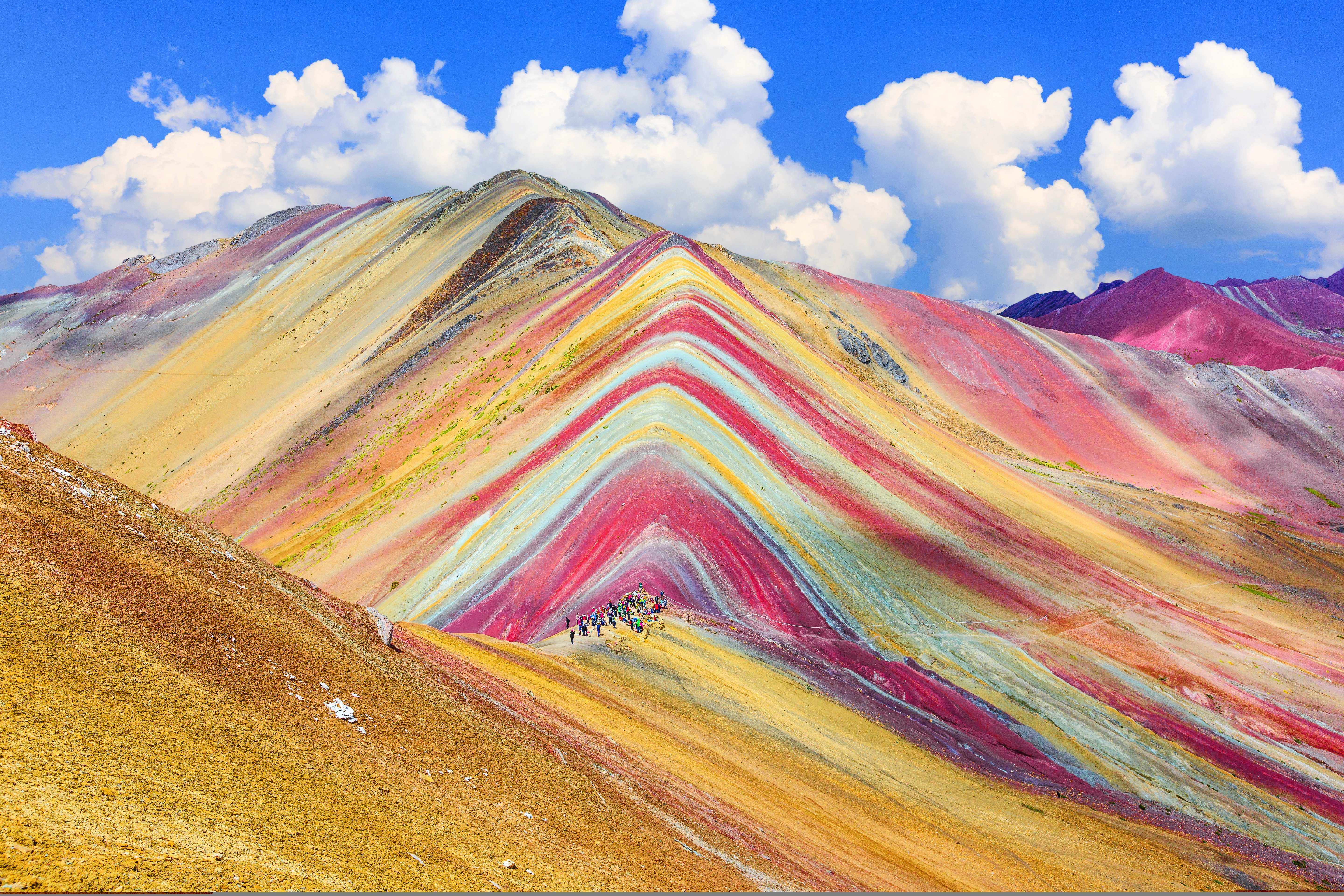 A wide-angle shot of Rainbow Mountain near Cuzco in Peru. The mountain's striations are brightly coloured, and a group of hikers has gathered to admire it. 