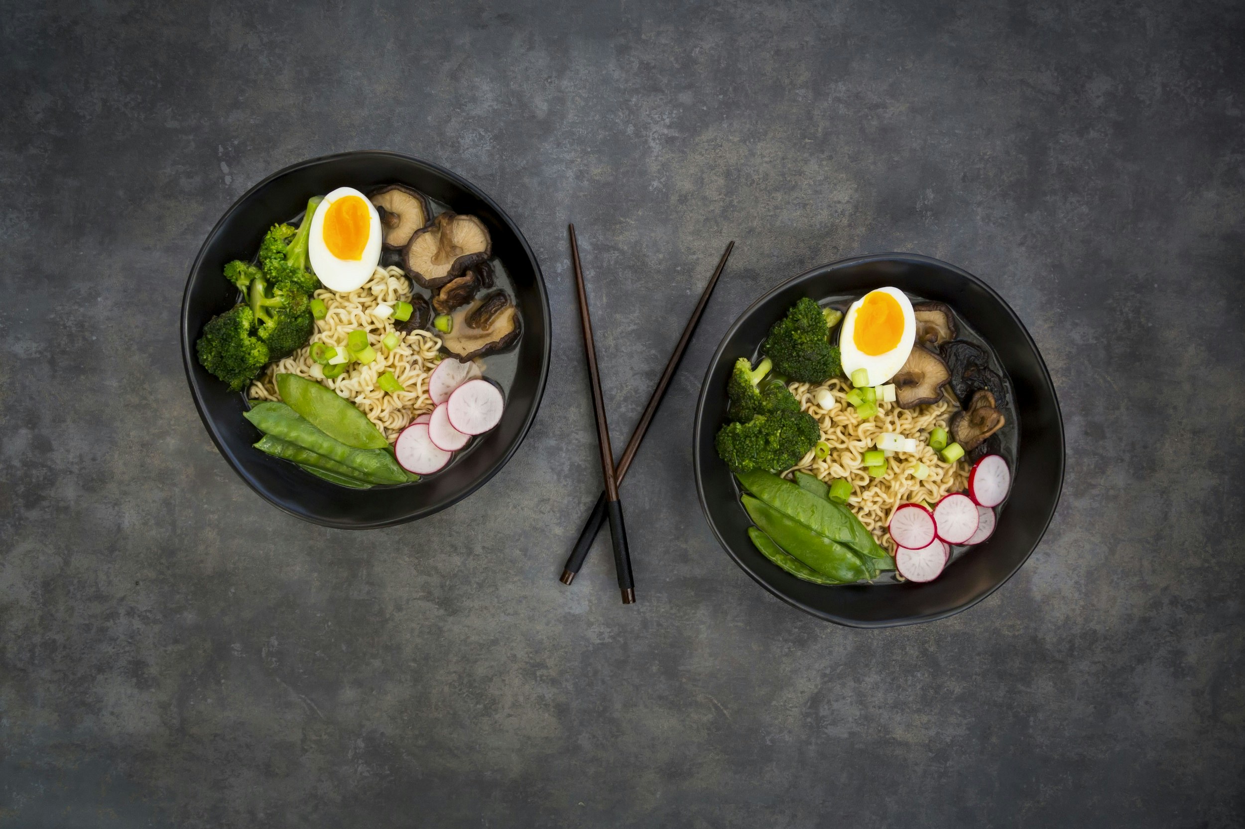 Two black bowls sit on a polished concrete counter, separated by dark brown chopsticks; within each bowl are neatly placed piles of ramen noodles, broccoli, sliced beets, pea pods, mushrooms and half a soft-boiled egg.