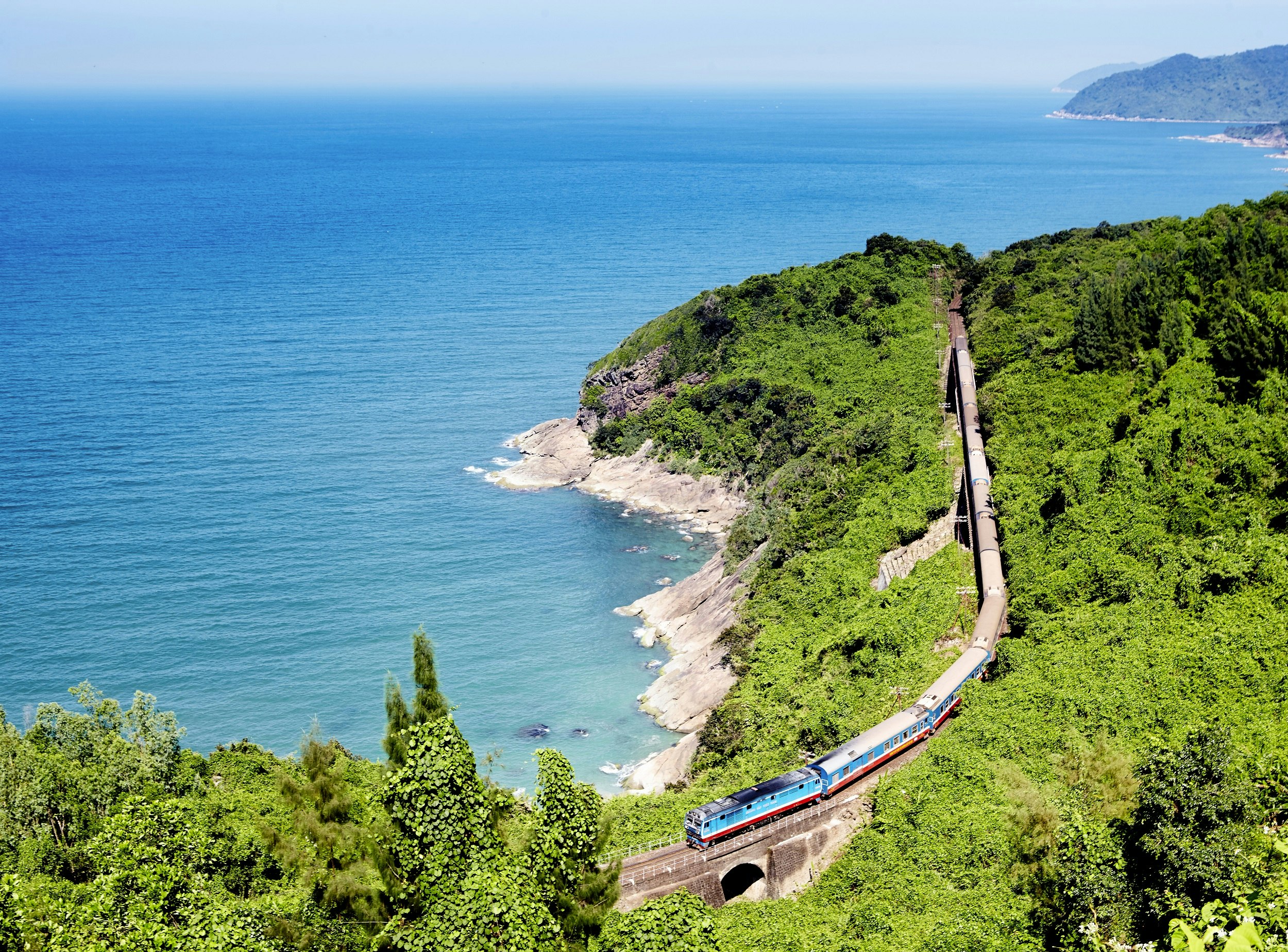 An aerial photograph of a train crossing a small bridge along a heavily forested section of coastline; a deep blue sea sits off the rocky shore that meanders in and out of the image.