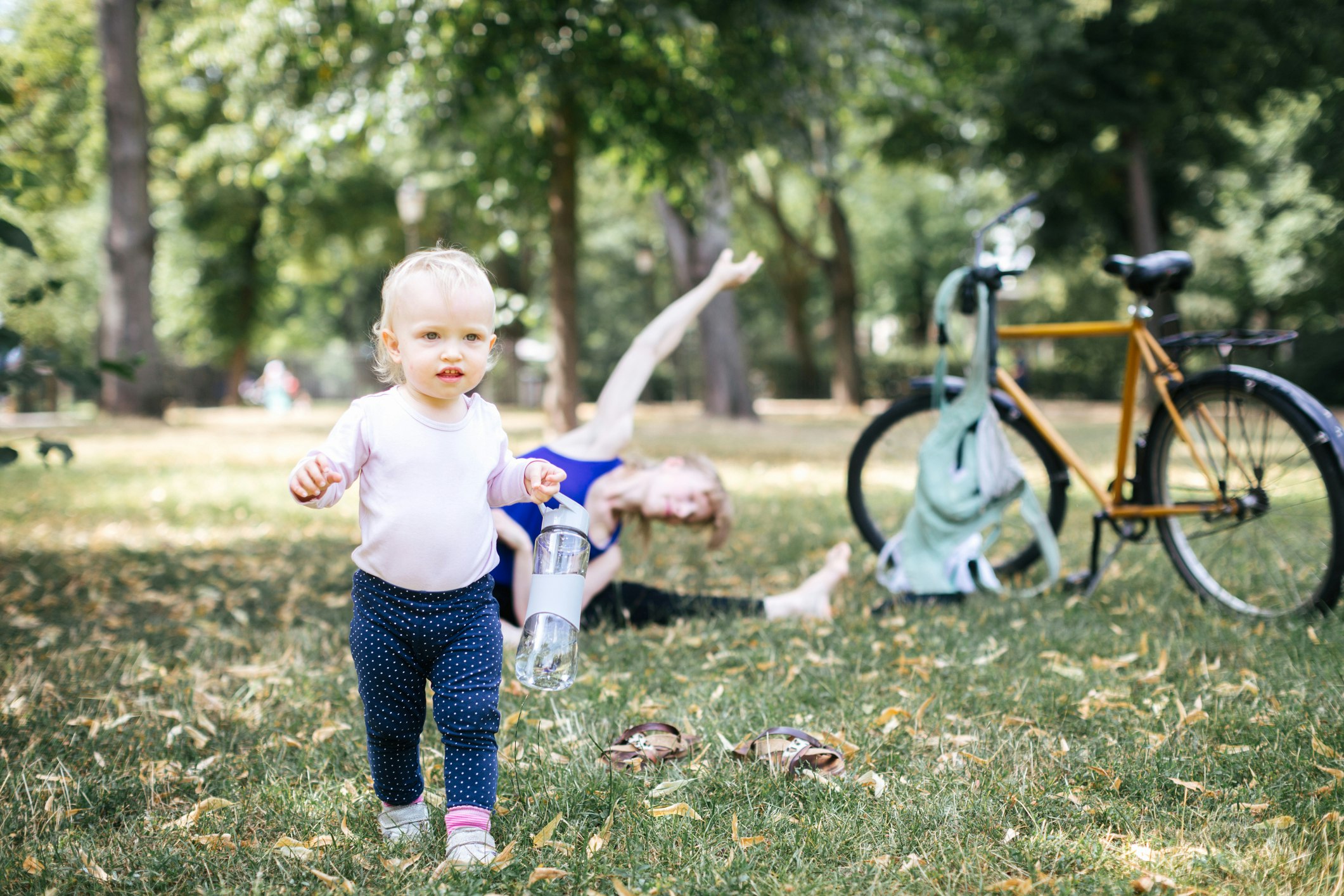 A tow-headed baby runs through a park with a reusable water bottle while her mother does yoga in the background in a purple top and black leggings. A yellow bicycle is parked nearby with a green bag hanging from the handlebars.