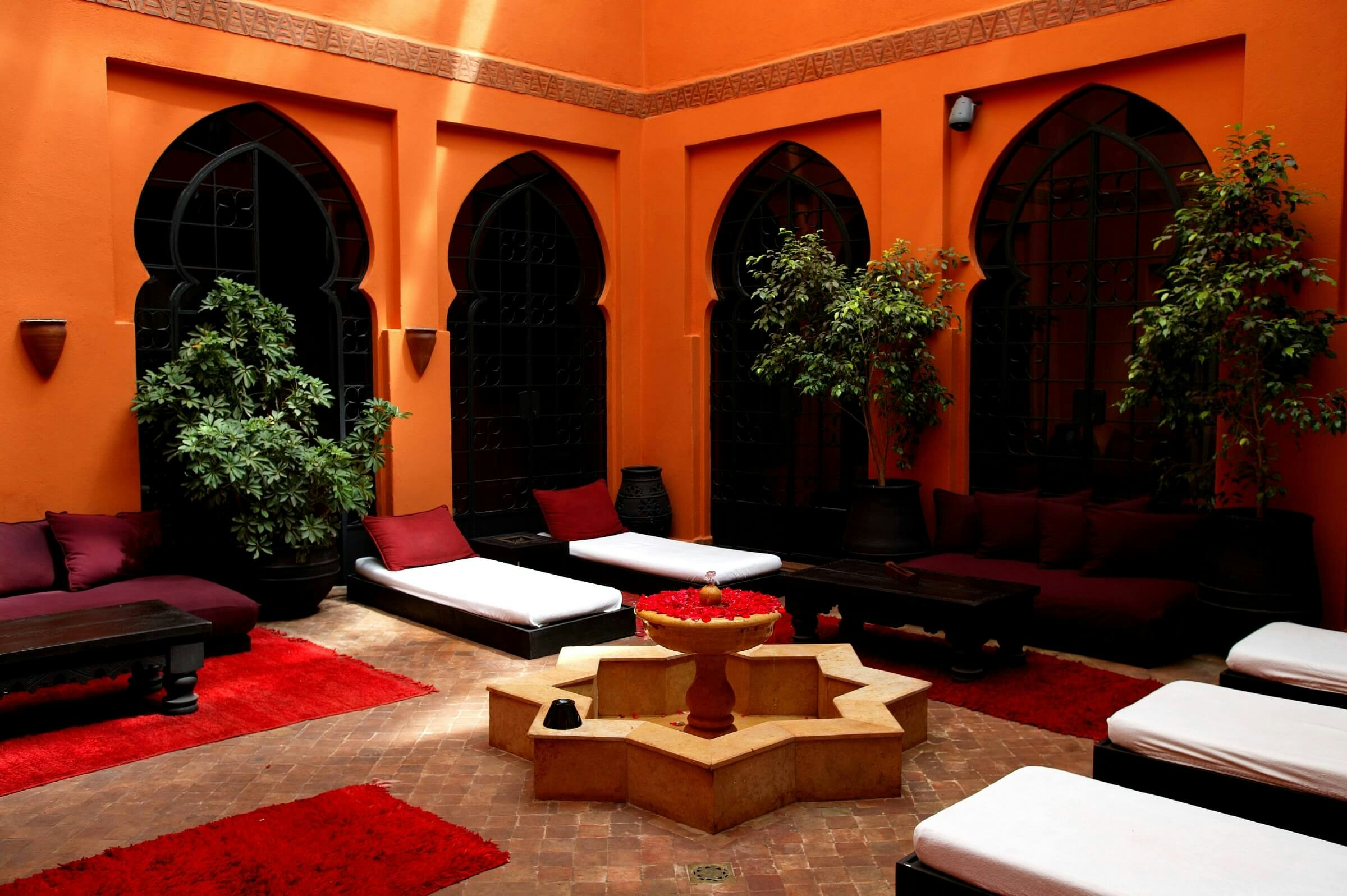 A riad with orange walls, a fountain in the middle of the room and loungers dotted around the room.