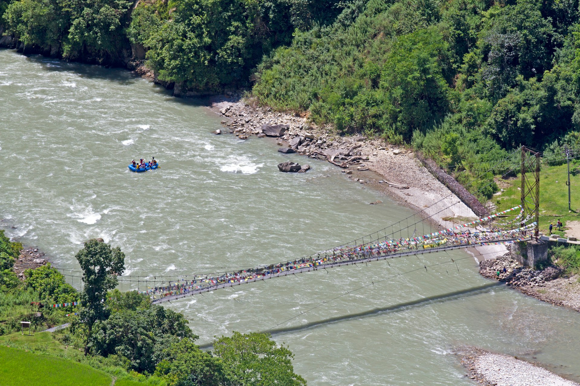 A blue river raft makes its way down a greenish-grey river that is lined with gravel banks and forests; they have just past beneath a prayer flag-covered footbridge that spans the river.