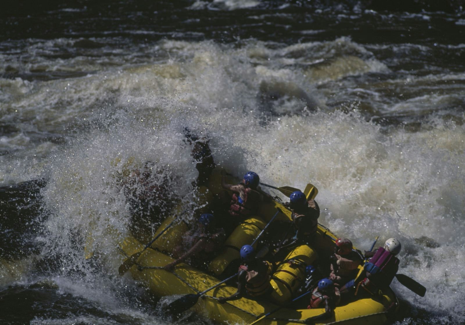 A river raft is deluged and almost turned over by an intense set of rapids on the Zambezi; most of the rafters have one hand on a paddle and the other firmly on the raft's safety line.
