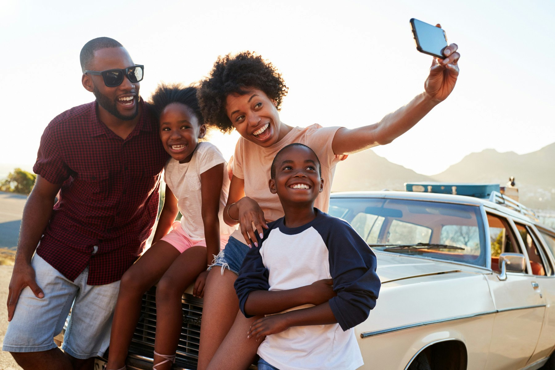 A young mom takes a selfie of a man and two children posing on the hood of a car.
